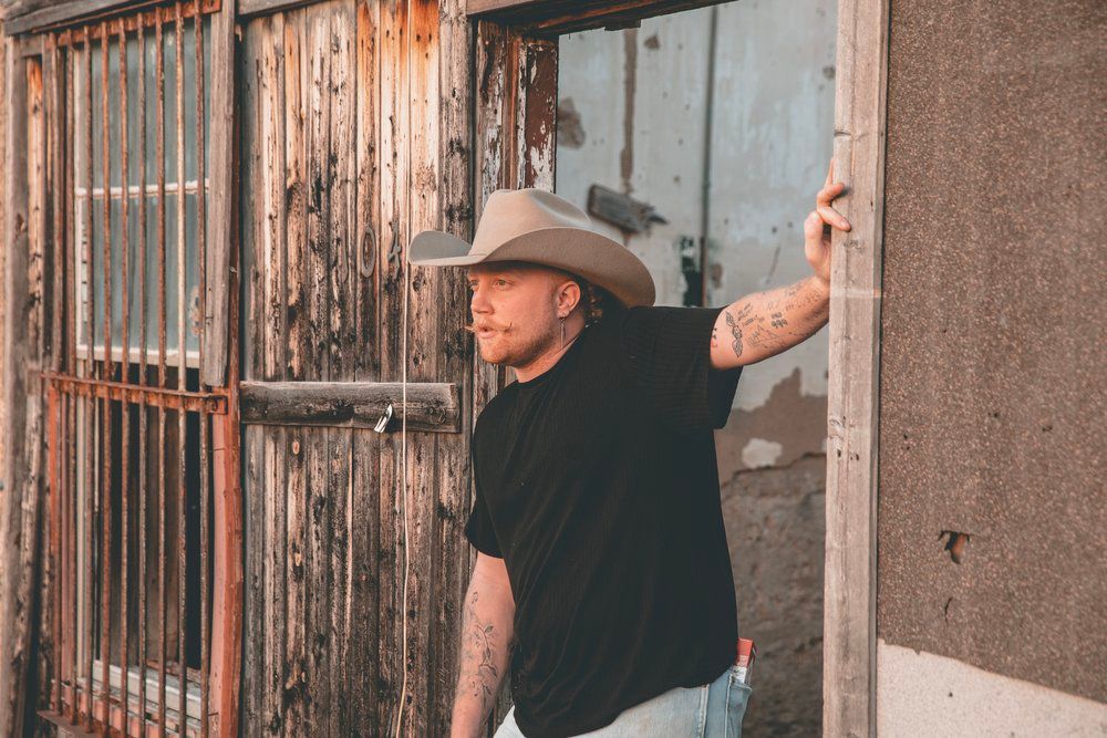 Person in black shirt and cowboy hat leans against weathered building wall with rustic windows.