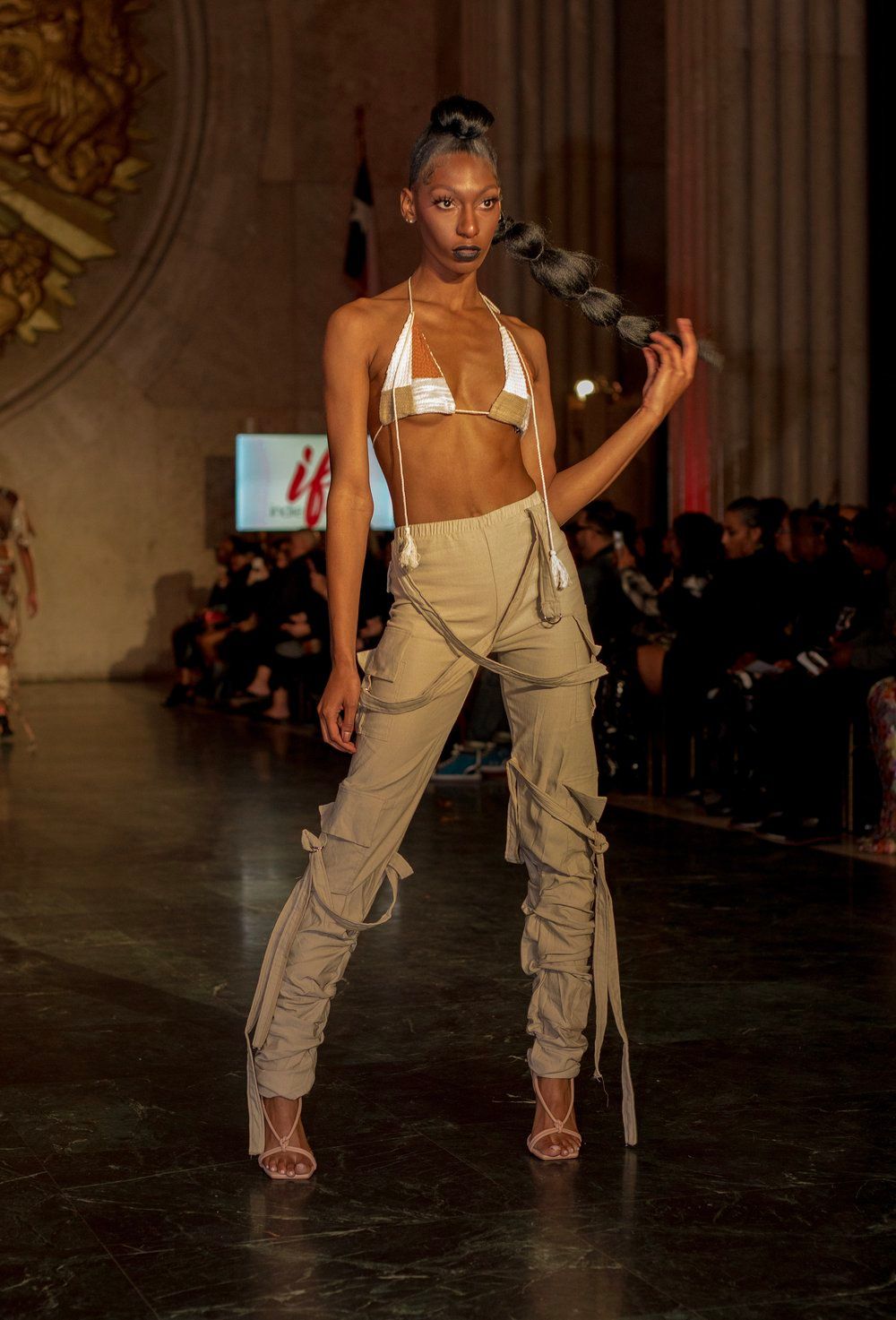 Fashion model walks runway in beige cargo pants and white bikini top during luxury fashion show presentation.