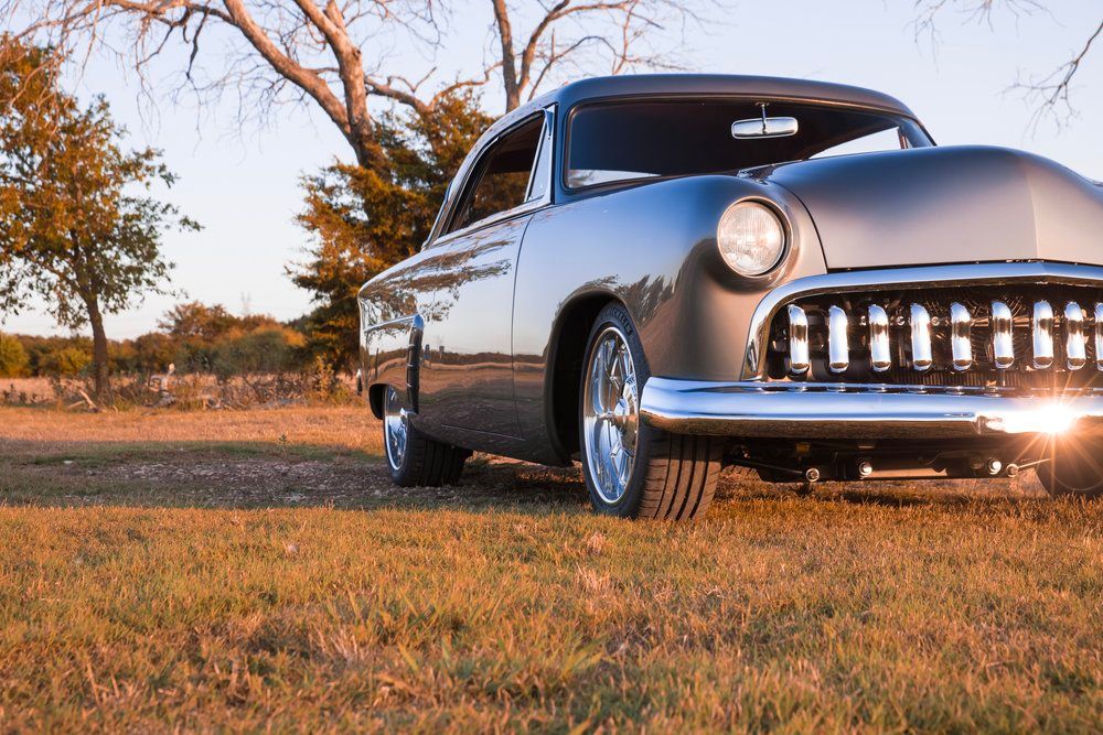 A classic silver hot rod with chrome trim sits in a grassy field during golden hour sunset.