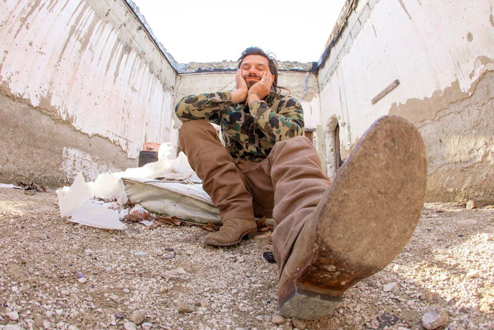 A person in military gear sits against a white wall, holding their head.