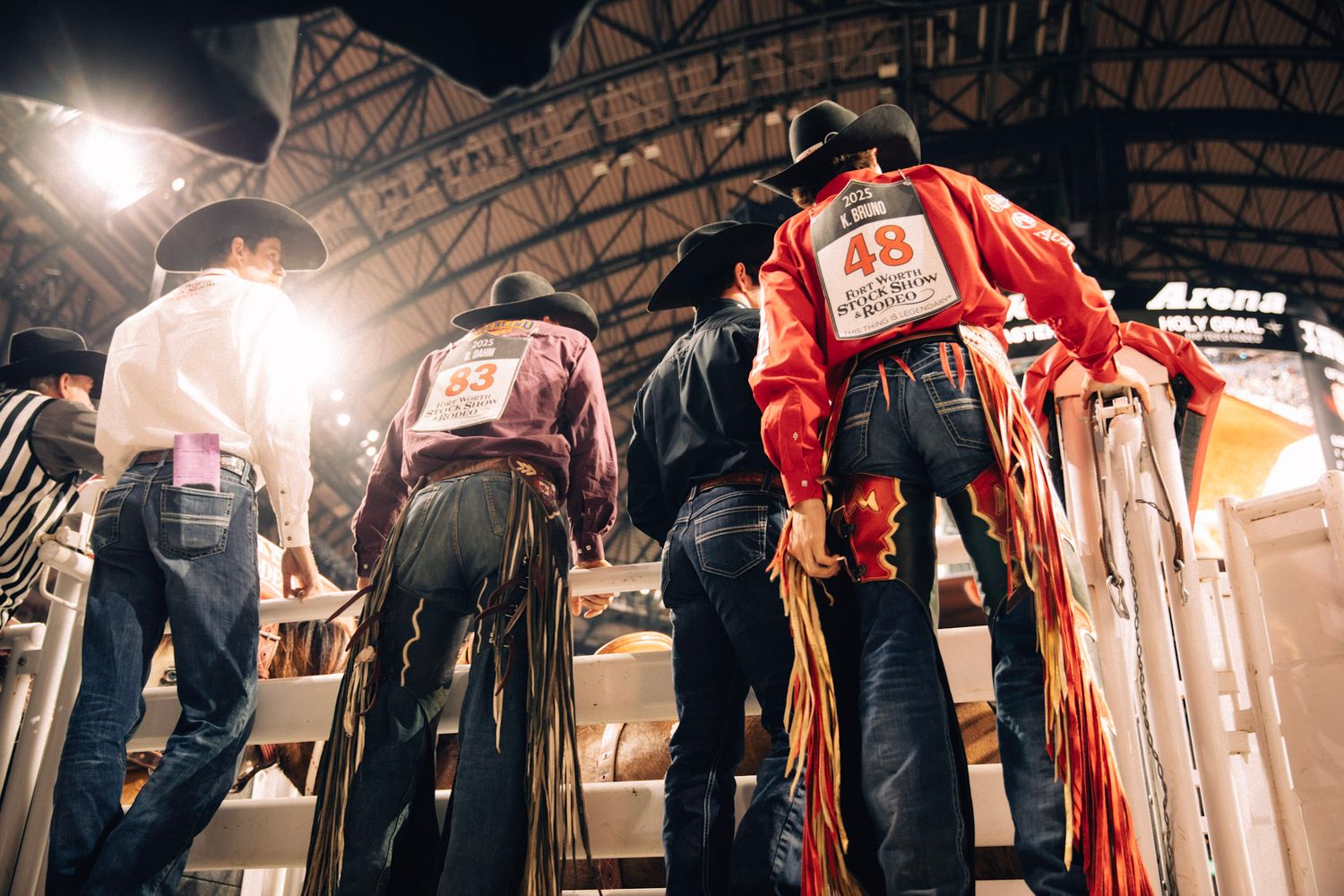 Low angle view of rodeo contestants in jeans and numbered vests under arena lights.