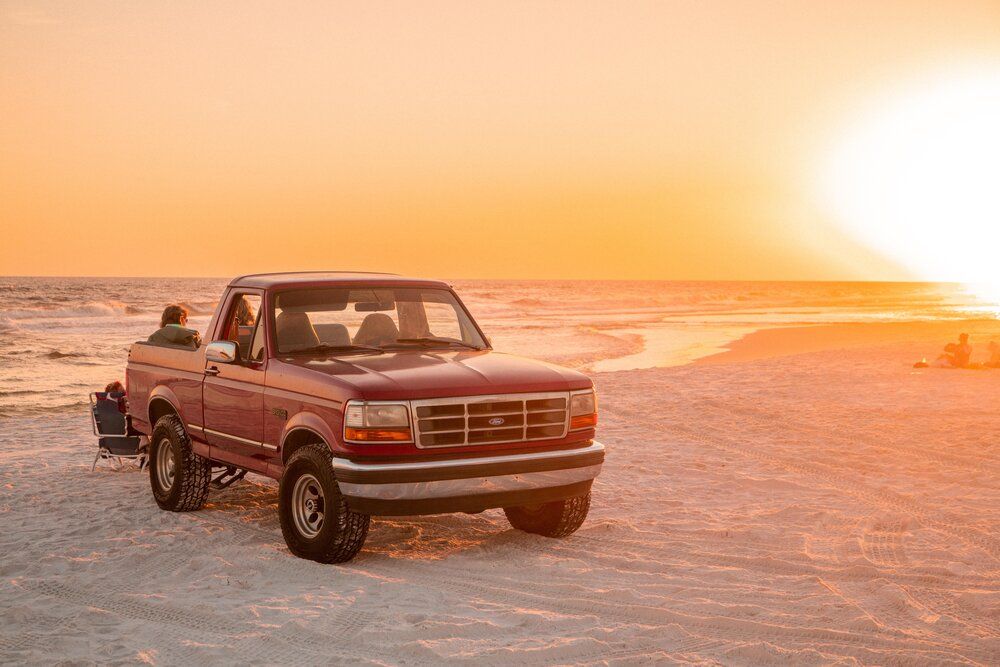 A series of pickup trucks silhouetted against a glowing orange sunset on a beach during golden hour.