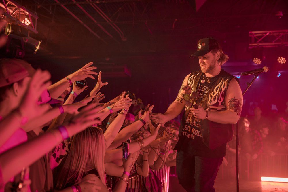 Performer Ernest reaches out to crowd of raised hands during energetic concert performance under red lighting.