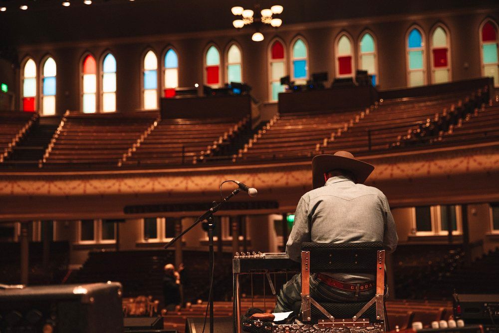 Musician sits alone in empty auditorium with stained glass windows during rehearsal or soundcheck.