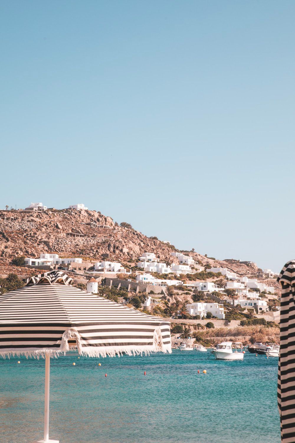 Beach umbrellas and turquoise waters along the coast of Mykonos with hillside buildings.