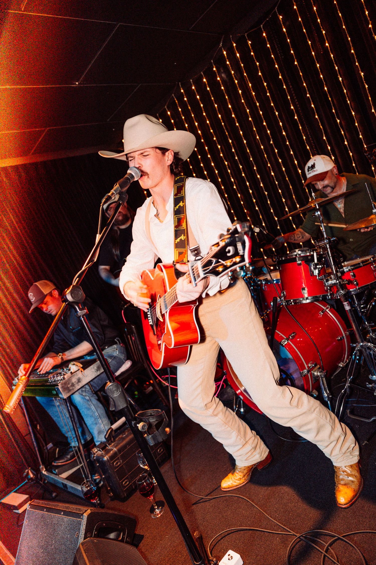 Country musician in white suit and cowboy hat plays red guitar on stage with band and string lights.