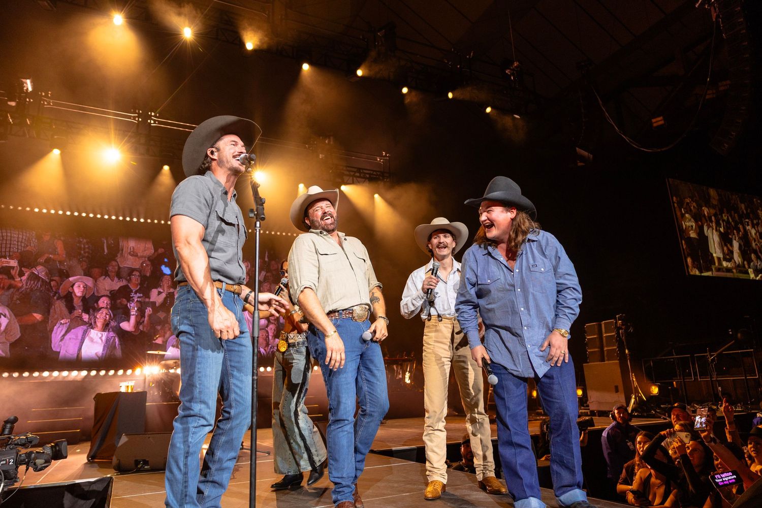 Four men in cowboy hats and jeans performing on a lit stage at a country music concert.