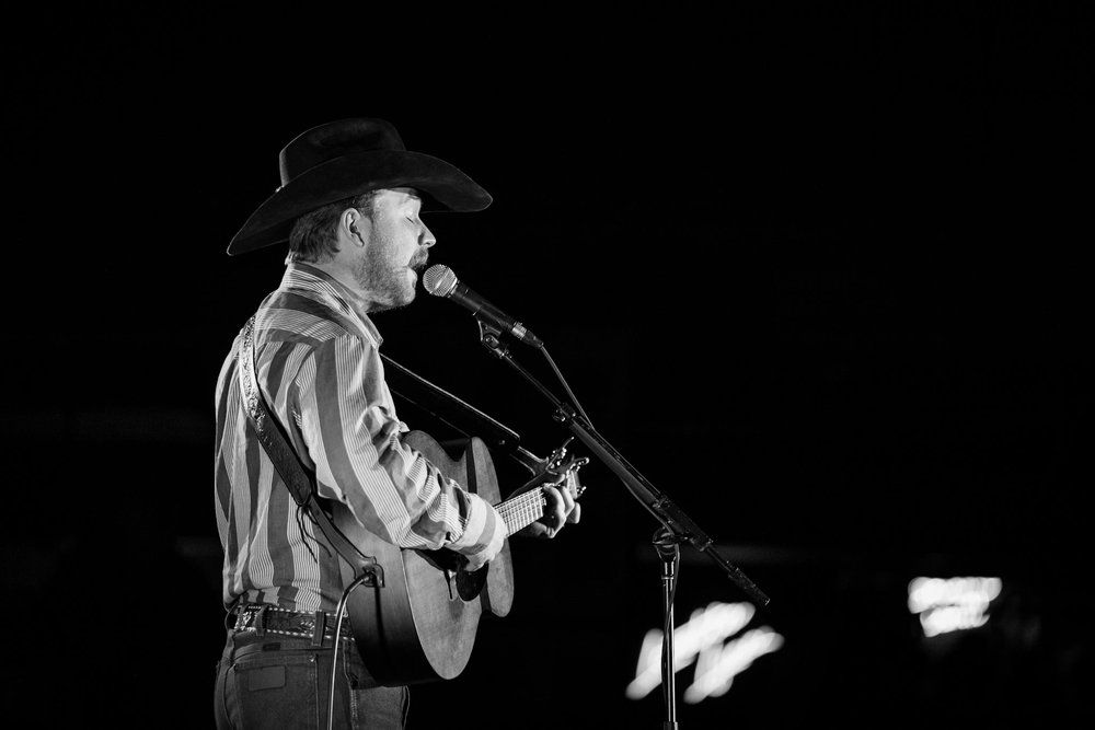 Black and white photo of a country performer Colter Wall singing into microphone while playing acoustic guitar on stage.
