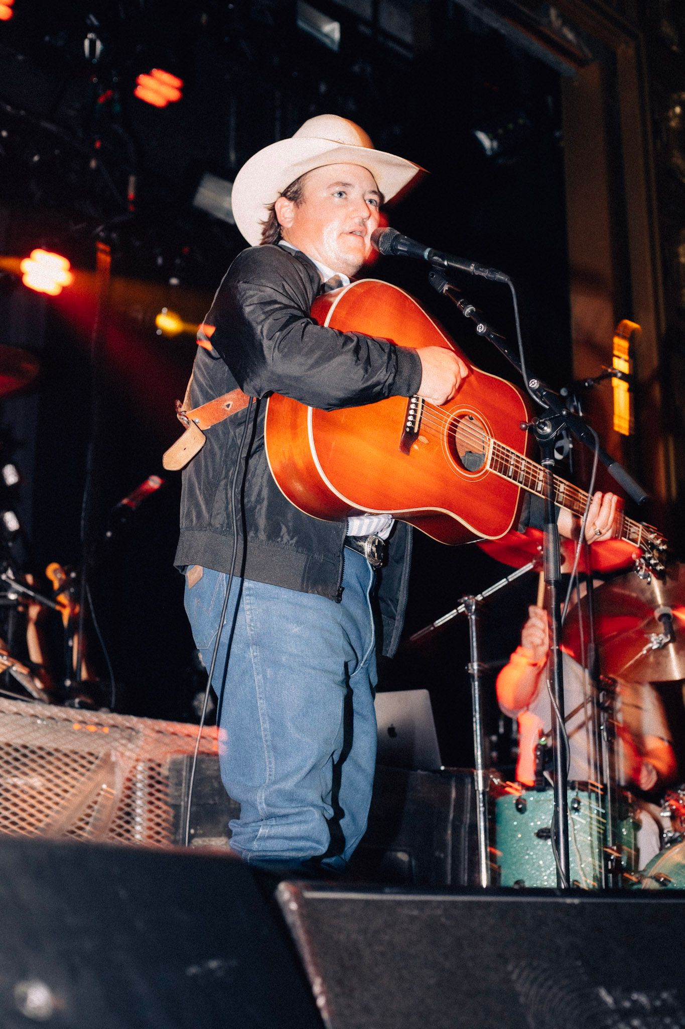 Jake Worthington performer in a cowboy hat and denim plays an acoustic guitar on stage under colored lighting during a concert.