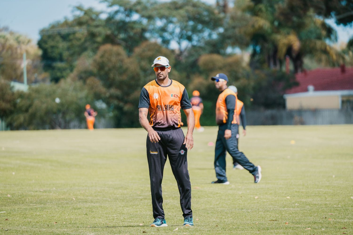 Cricket player in orange and black uniform standing on green field during a sunny outdoor match.
