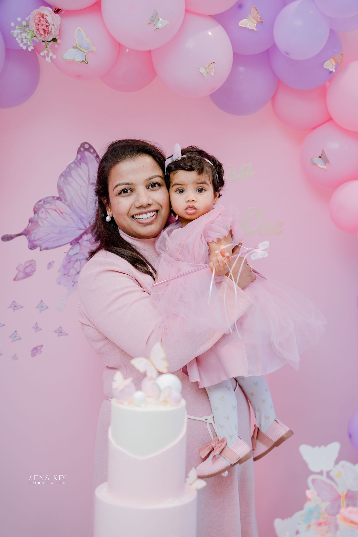 Mother holding baby daughter in pink outfits in front of butterfly and balloon birthday party backdrop.