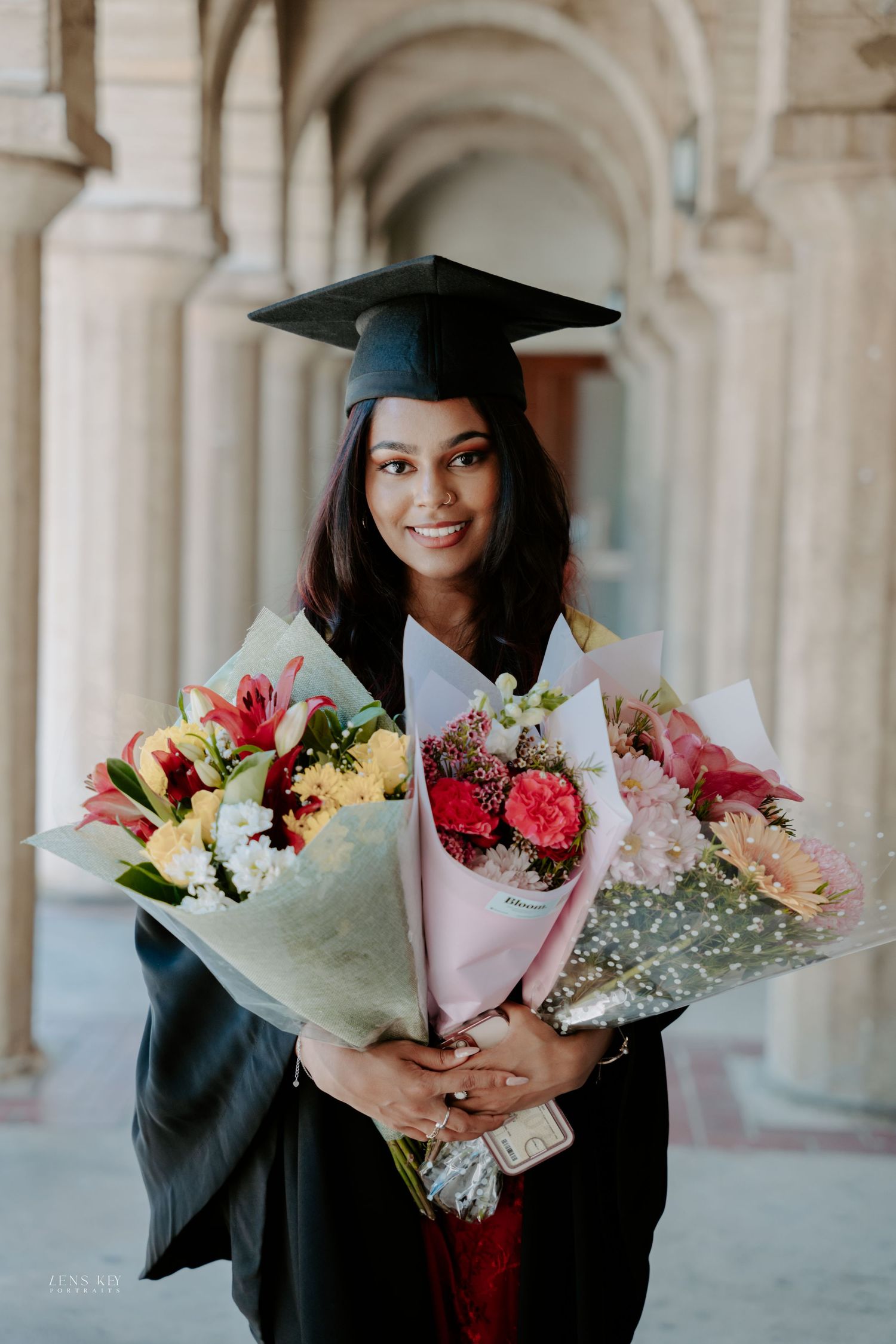 Happy graduate in cap and gown holds two large colorful flower bouquets under stone arches.