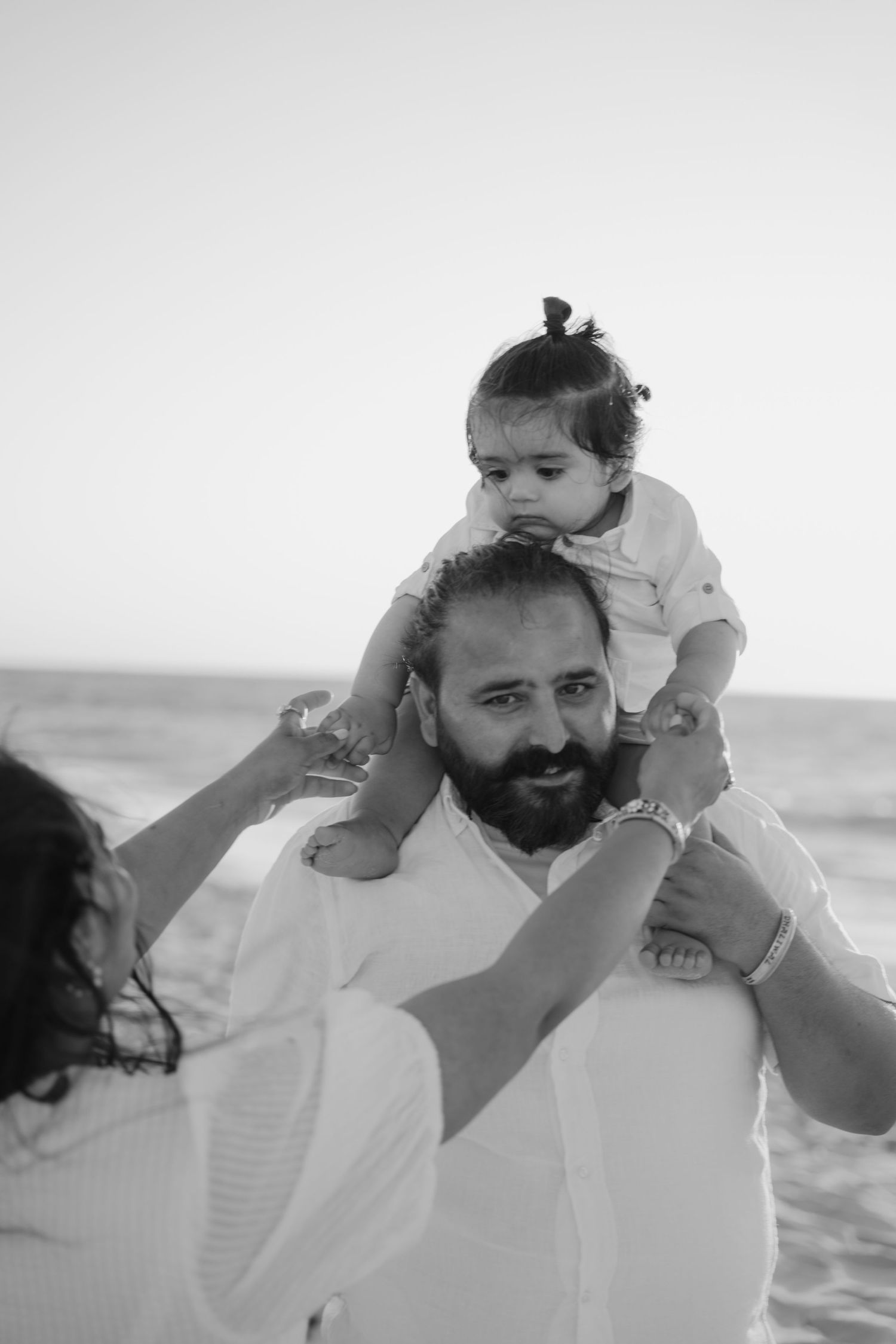Black and white photo of bearded father carrying baby on shoulders at the beach.