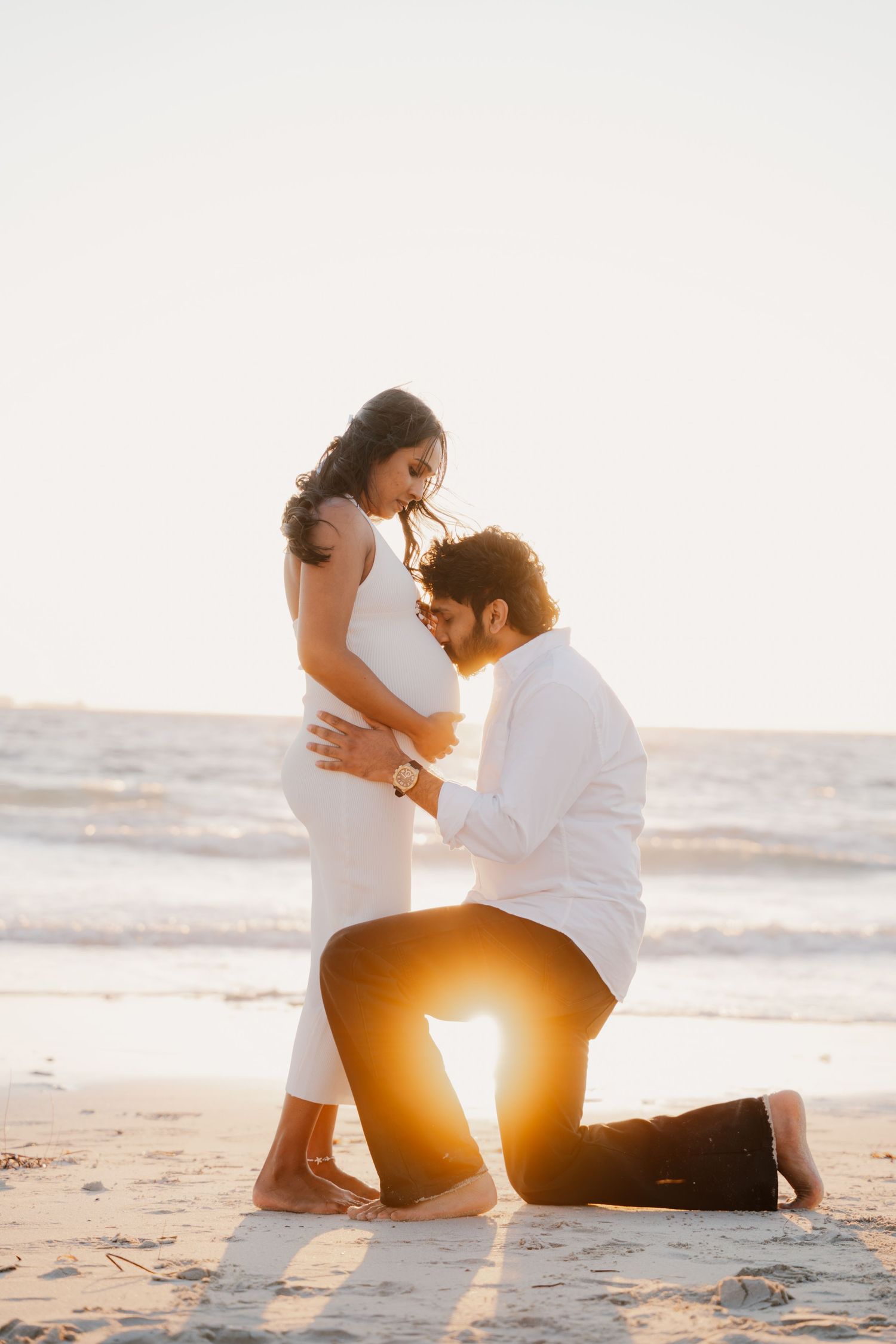 Man kneeling before pregnant woman on beach at golden sunset in a tender romantic moment.