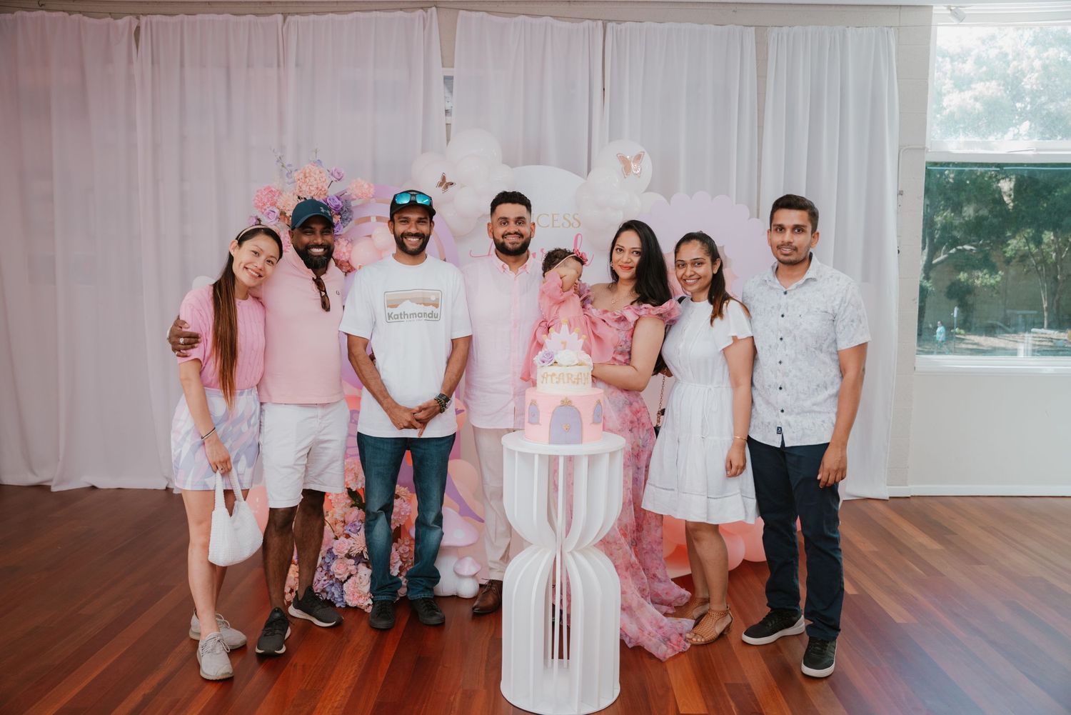 Group of seven guests poses with pink butterfly first birthday cake at celebration venue.