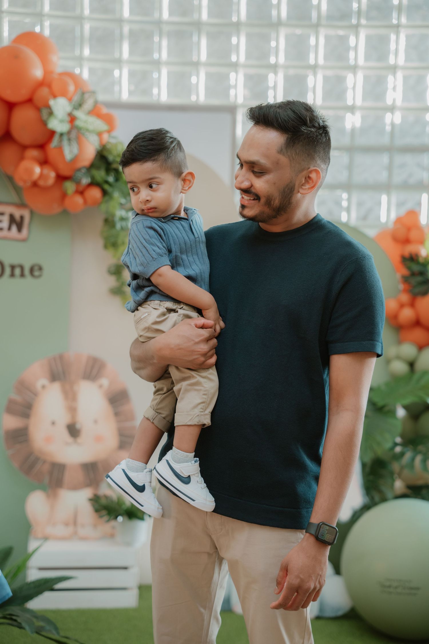 A man smiles while holding a toddler at a safari-themed first birthday party with orange and green balloon decor.