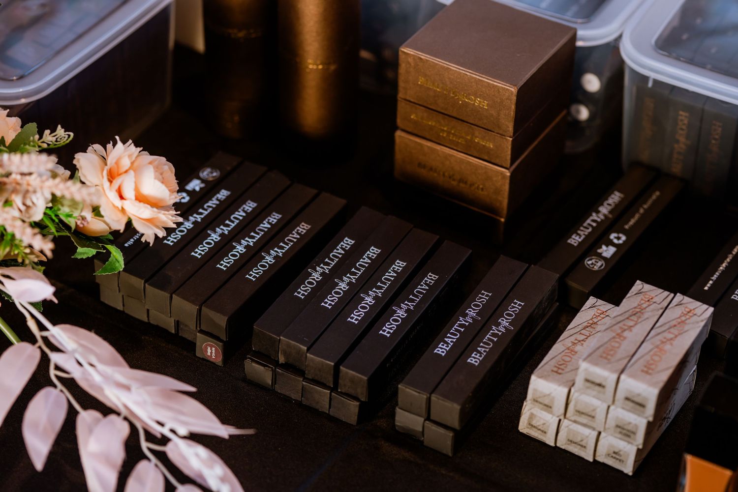 Close-up of stacked highlighter and beauty product boxes on a dark surface with pink flowers.