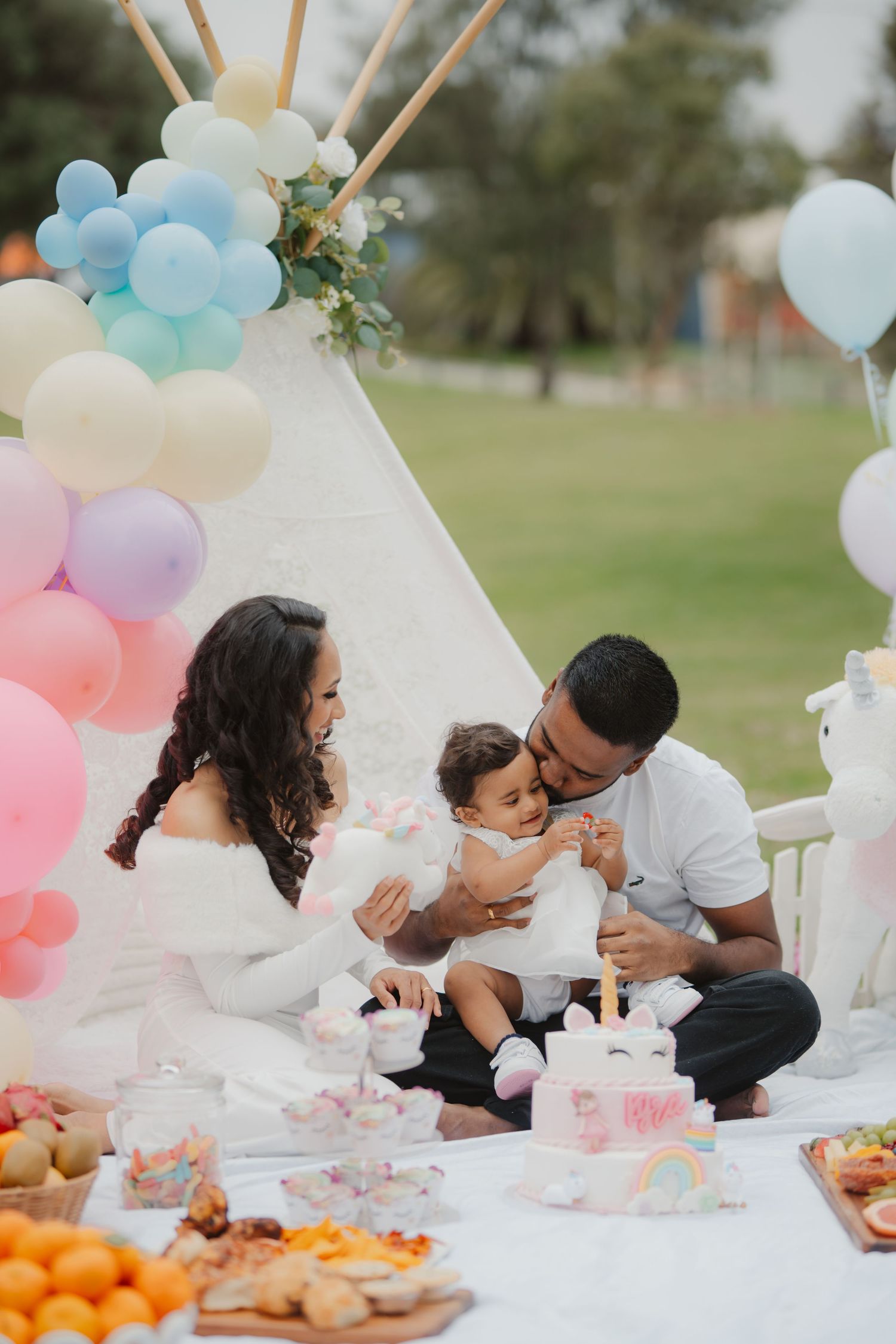 Parents and baby enjoying a unicorn themed first birthday picnic with cake and pastel balloons.