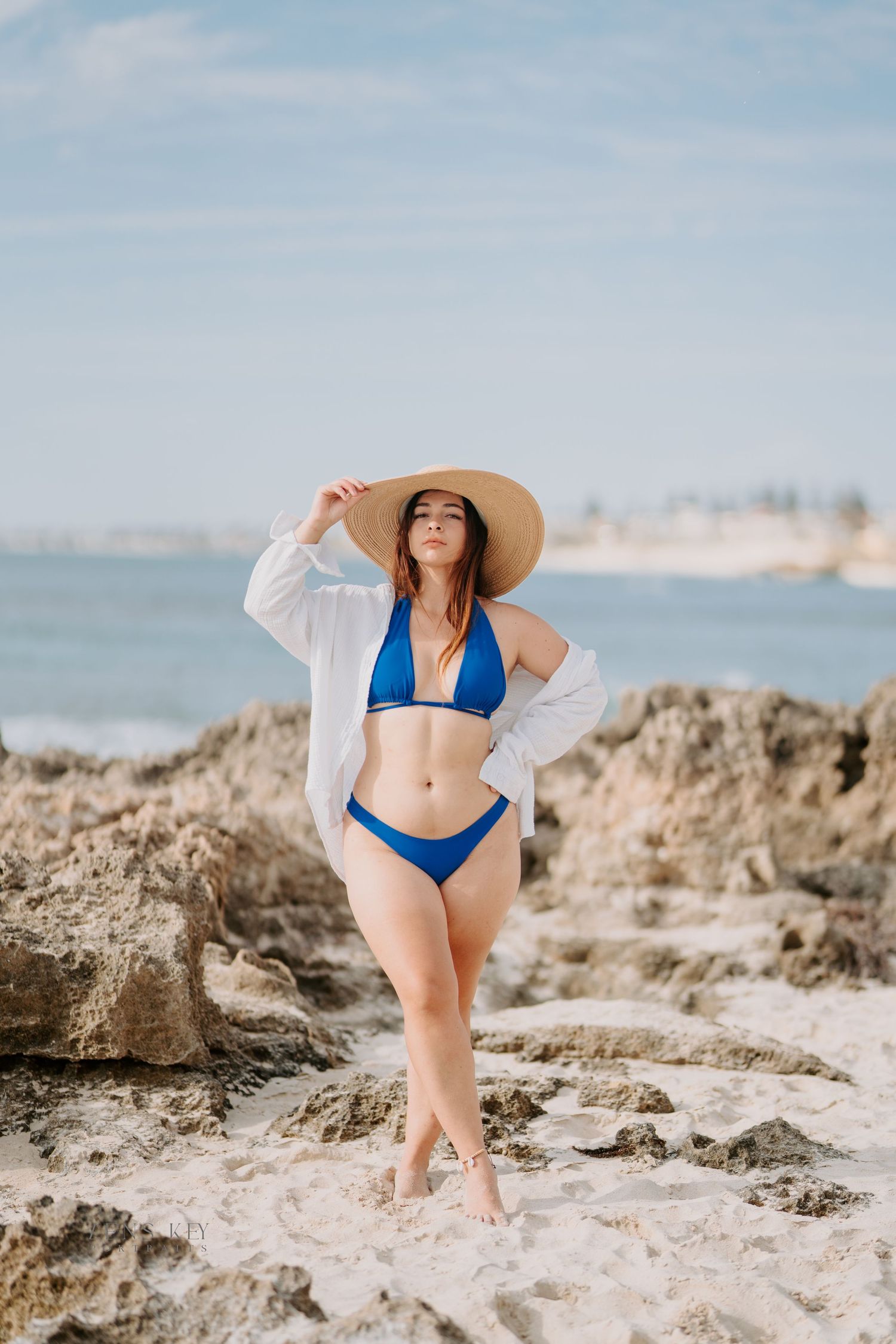 A woman in blue bikini and white shirt holds a wide-brim straw hat while posing on rocky beach.