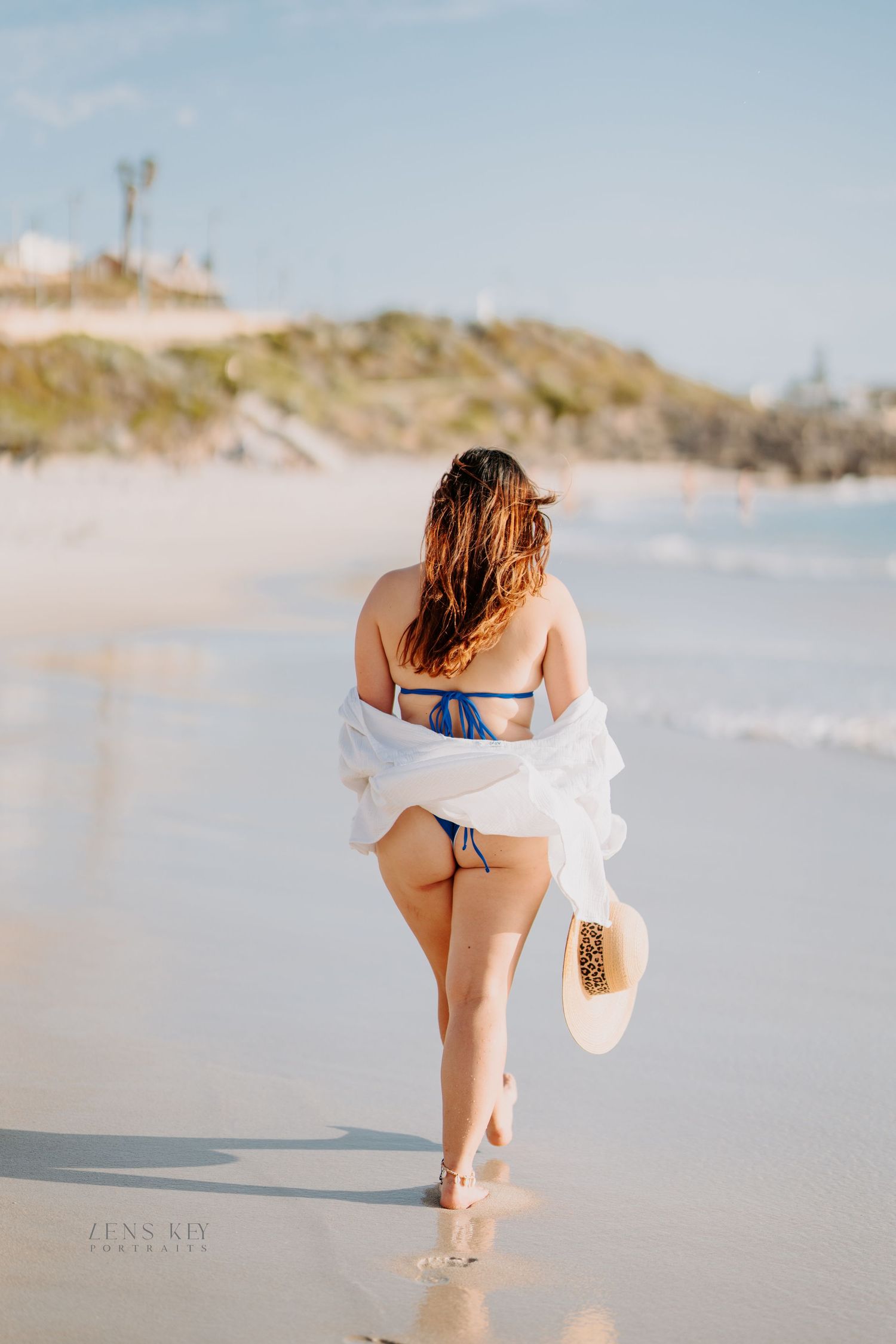 A woman in a blue bikini and white shirt walks along a sandy beach holding a straw hat.