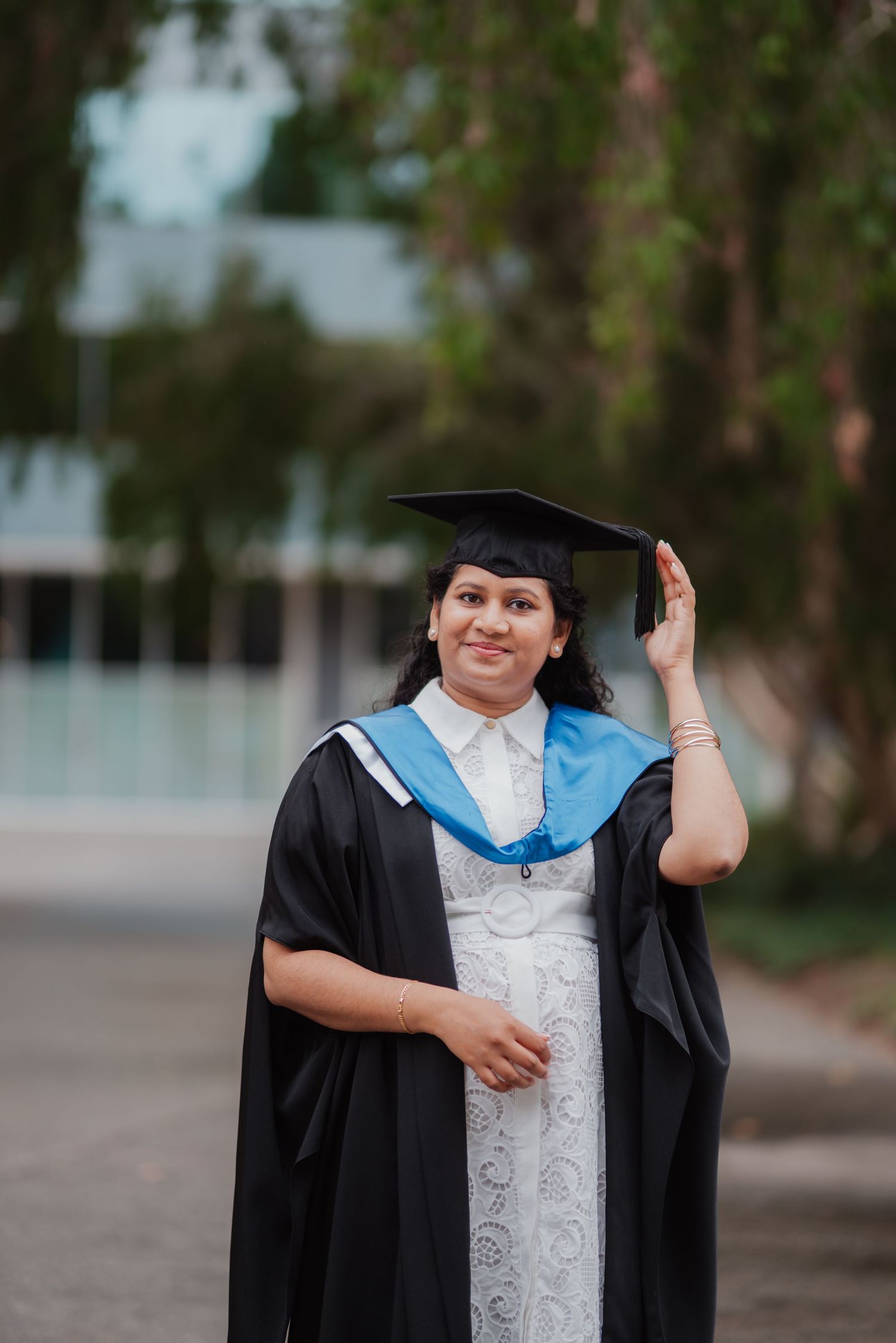 Happy graduate in black cap and gown adjusts mortarboard while smiling outdoors on campus.