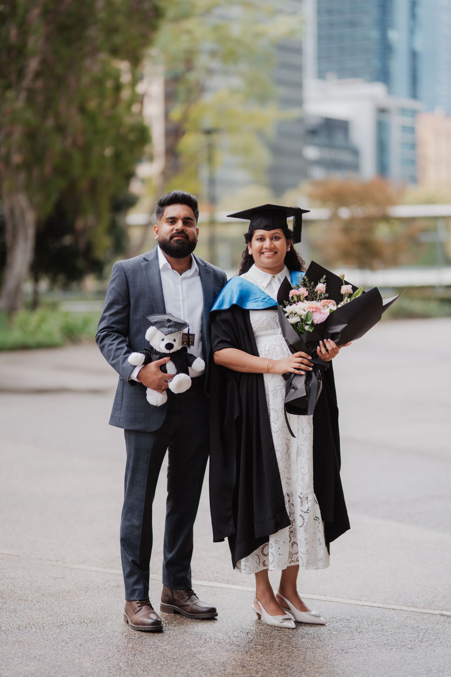 Male and female graduate couple pose outdoors holding flowers and graduation teddy bear.