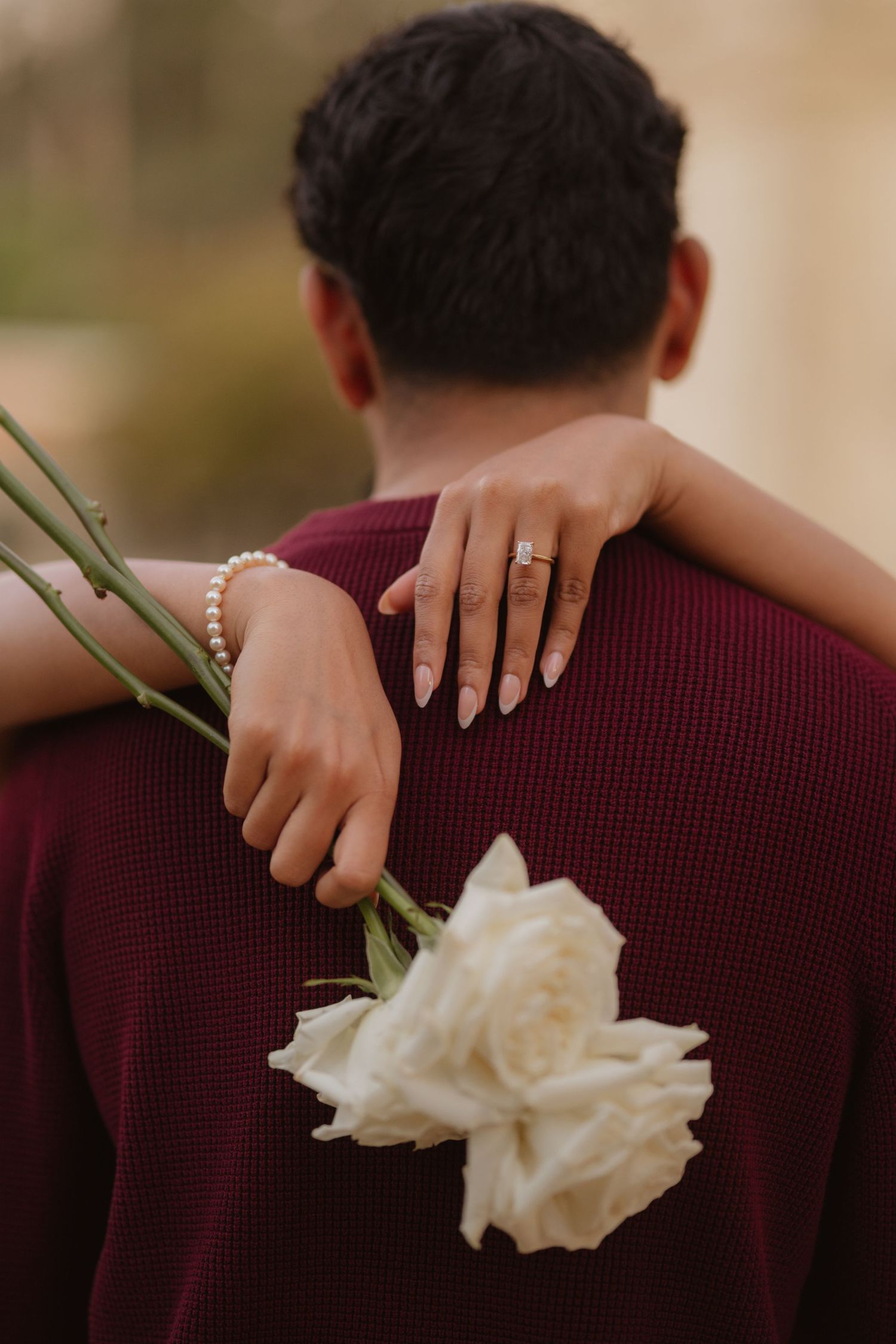 A woman embraces a man from behind wearing a burgundy sweater, holding white roses with an engagement ring visible.