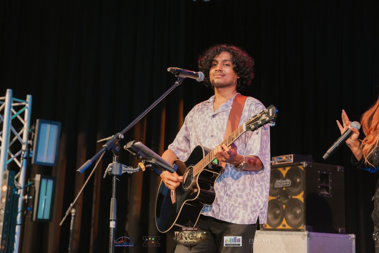 Young guitarist in floral shirt smiling while playing acoustic guitar on stage.