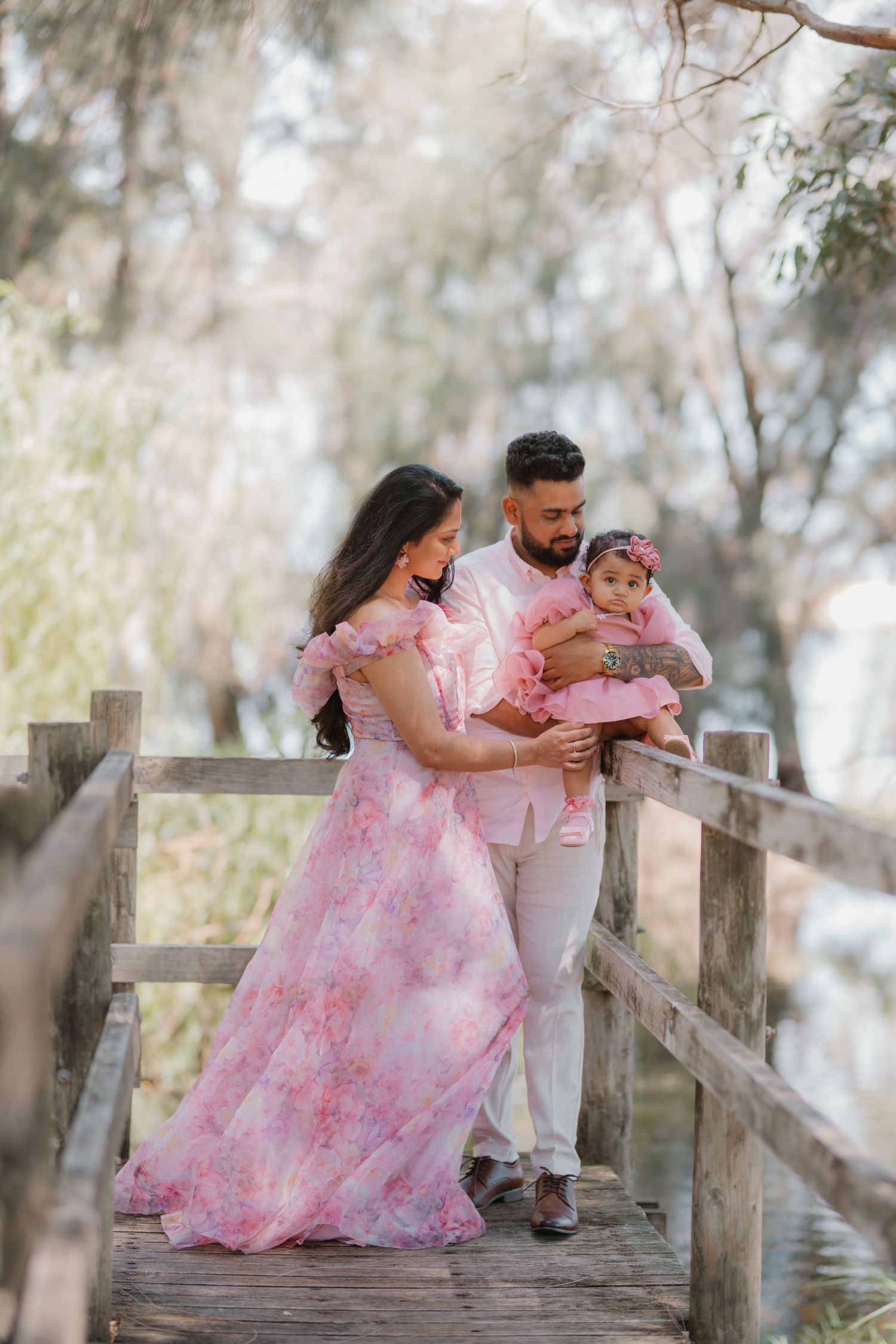 Family in matching pink outfits poses on wooden bridge, mother in floral gown holding baby daughter.
