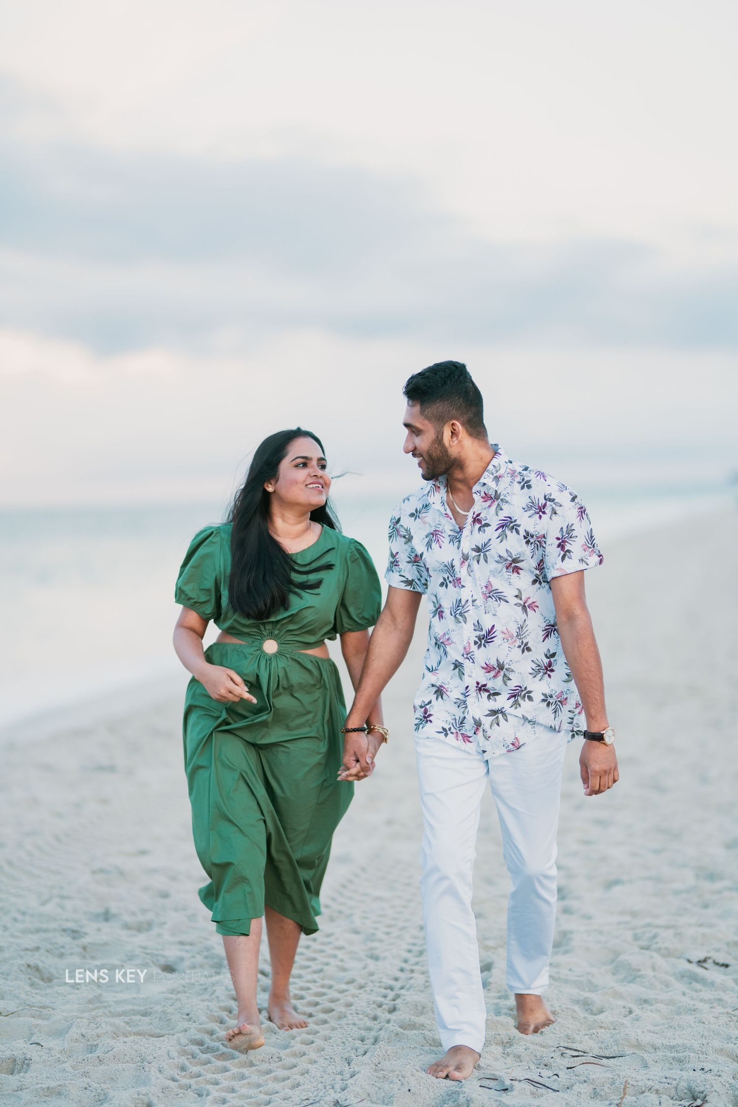Couple holding hands and walking along a sandy beach, woman in green dress and man in floral shirt.