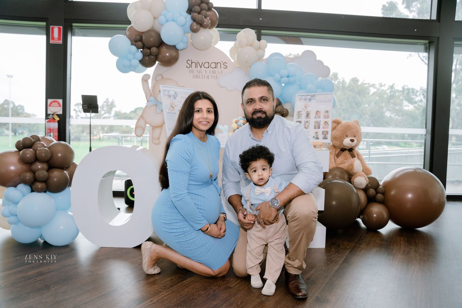 Family posing at a baby's first birthday party with blue and brown balloon decorations.