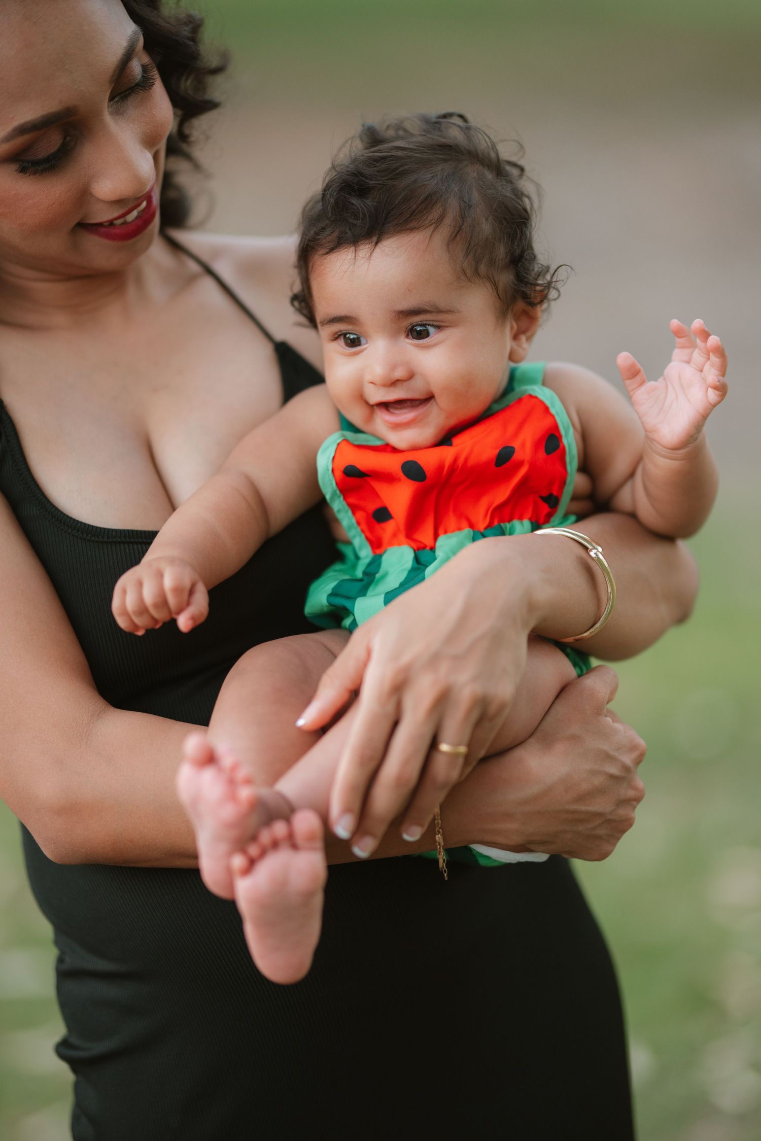 A smiling baby in a watermelon outfit being held by a woman in a black dress outdoors.