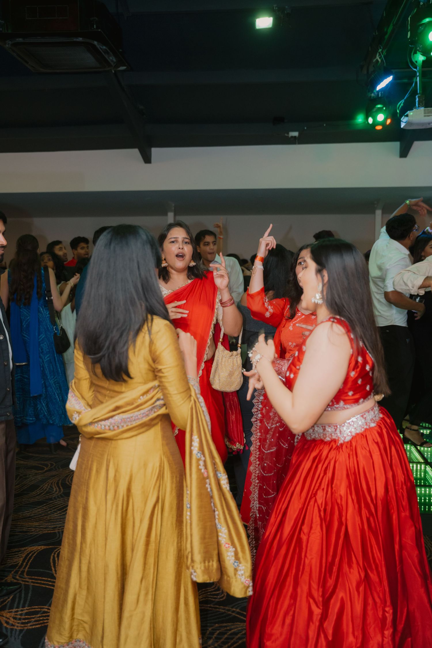 Women in vibrant red and gold Indian lehengas dancing joyfully at wedding reception party.