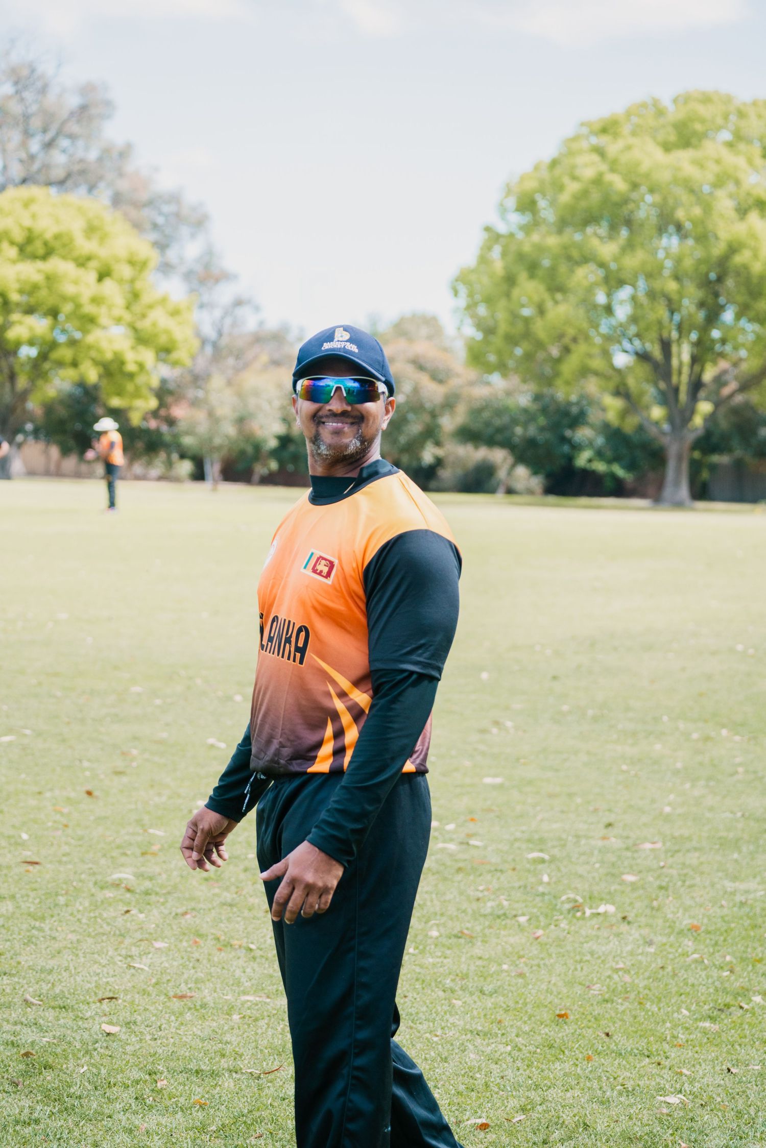Smiling Sri Lankan cricketer in orange jersey and sunglasses on green field.