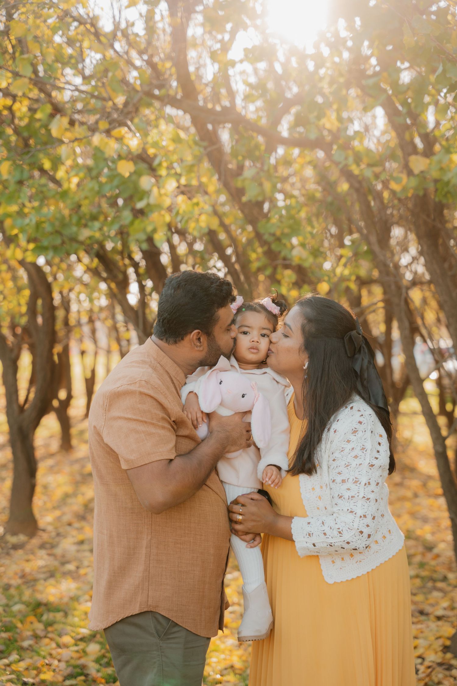 Parents kiss their baby girl on the cheeks in a golden autumn forest bathed in warm sunlight.