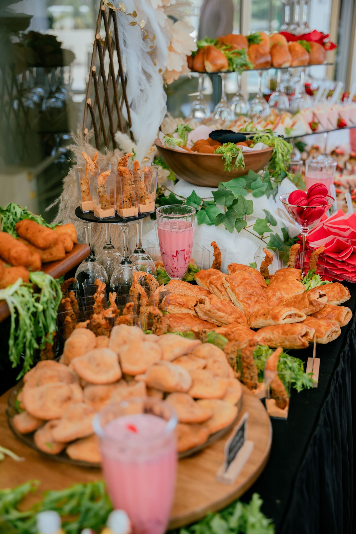 Elegant birthday party food spread with golden pastries, fried appetizers, pink drinks, and fresh greens on wooden boards.