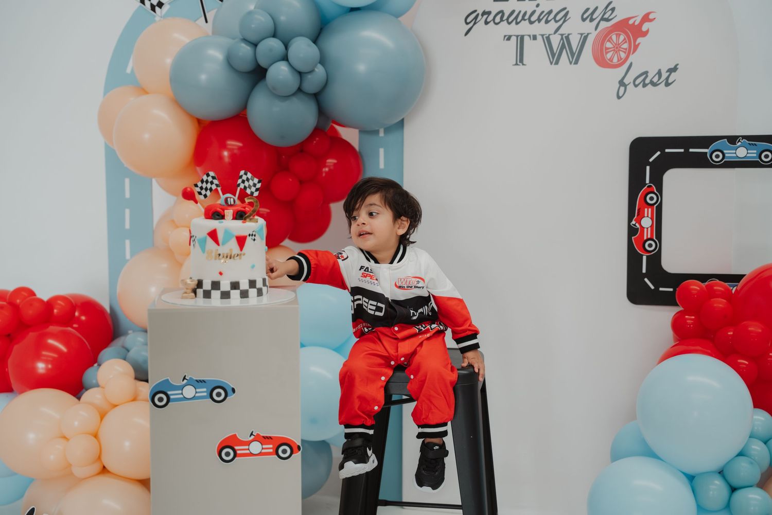 A toddler in a racing suit sits at his race car TWO fast second birthday party with balloon decor.