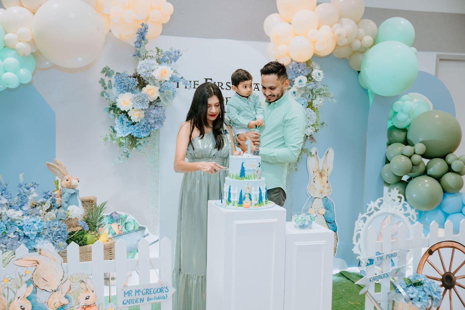 Parents hold their child while admiring a tall cake at a blue Peter Rabbit birthday party.