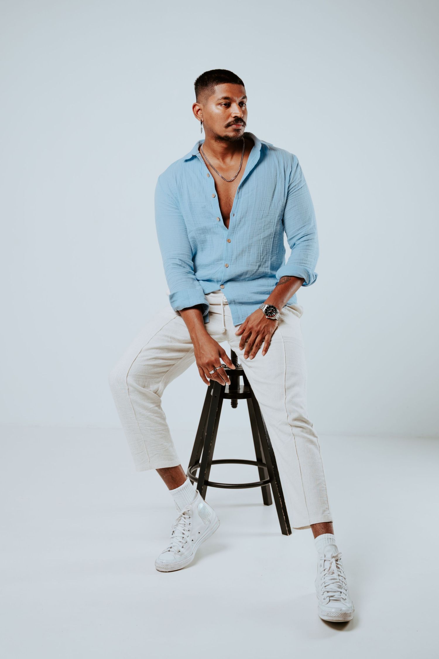 Man in blue linen shirt and white pants sits relaxed on dark wooden stool in studio.