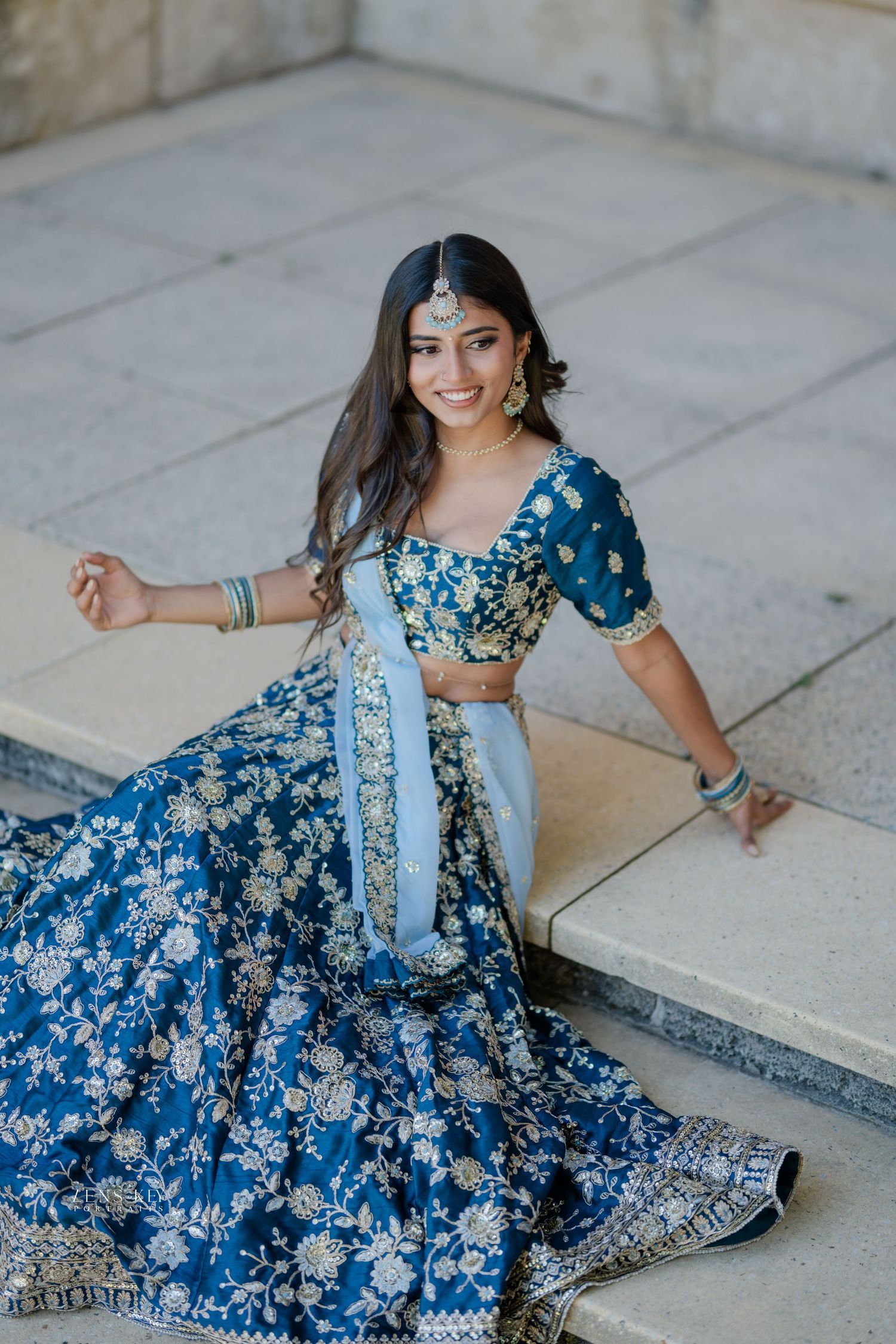 Smiling woman in blue floral embroidered lehenga choli seated on stone steps outdoors.