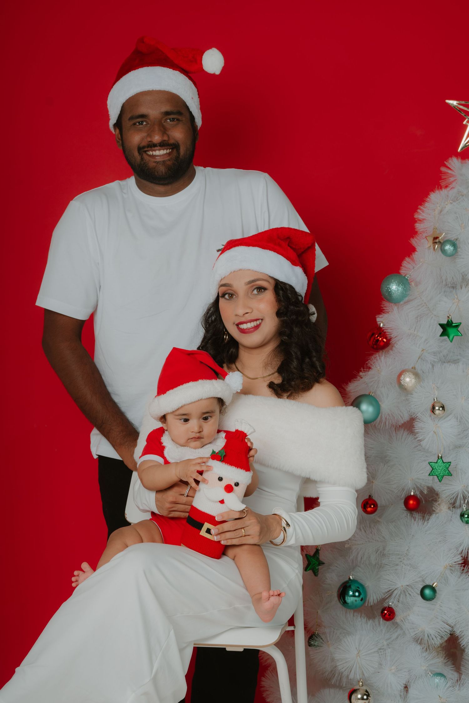 Family of three wearing Santa hats posing beside a white Christmas tree against a bold red background.