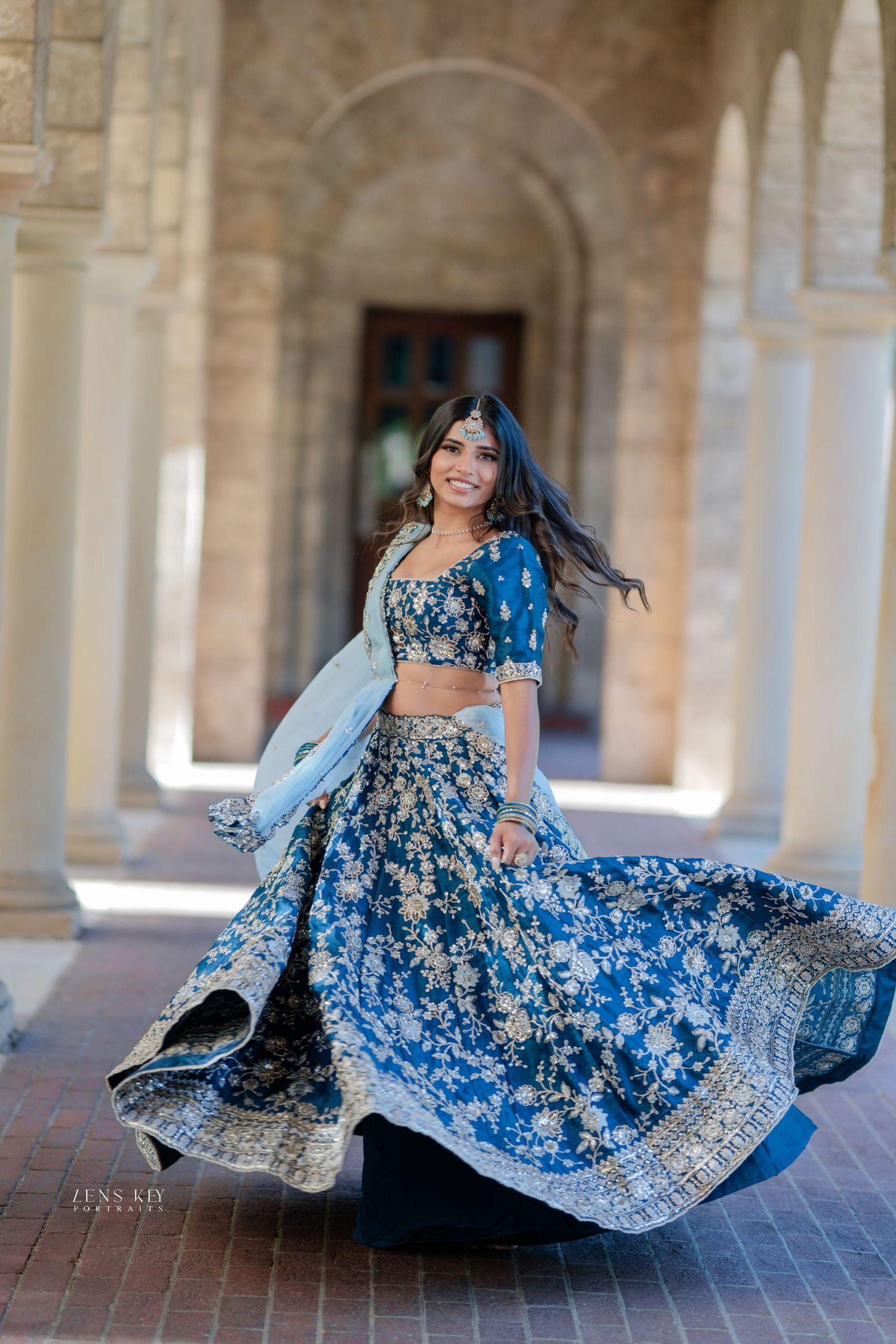 Woman twirling in blue floral embroidered lehenga choli in elegant columned corridor.
