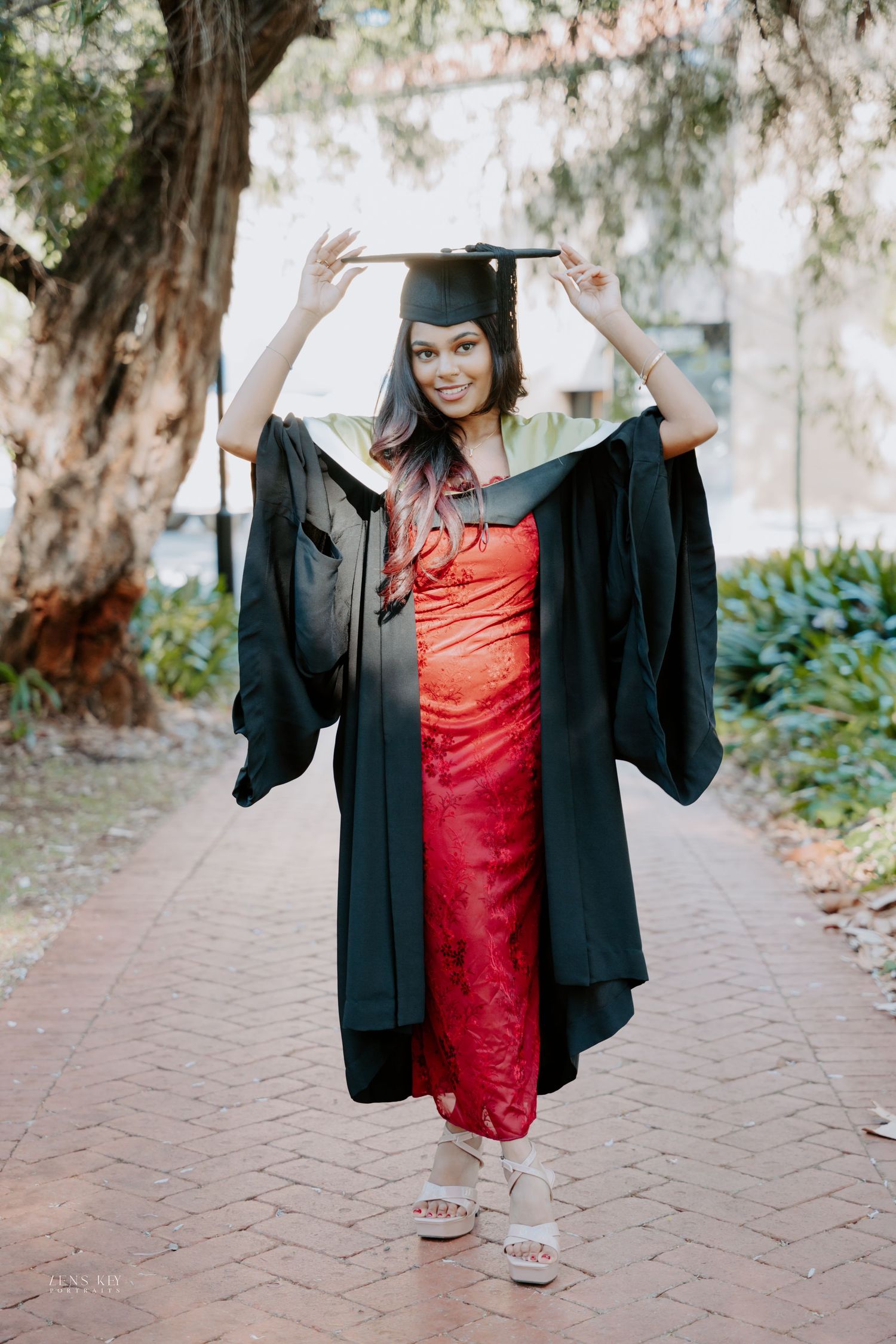 Smiling graduate in black cap and gown over red dress poses on brick pathway outdoors.