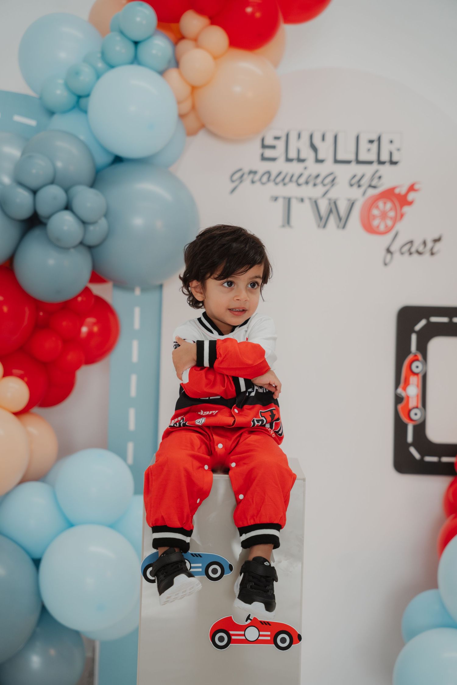 A toddler in a red racing suit sits on a white pedestal at a TWO fast race car birthday party.
