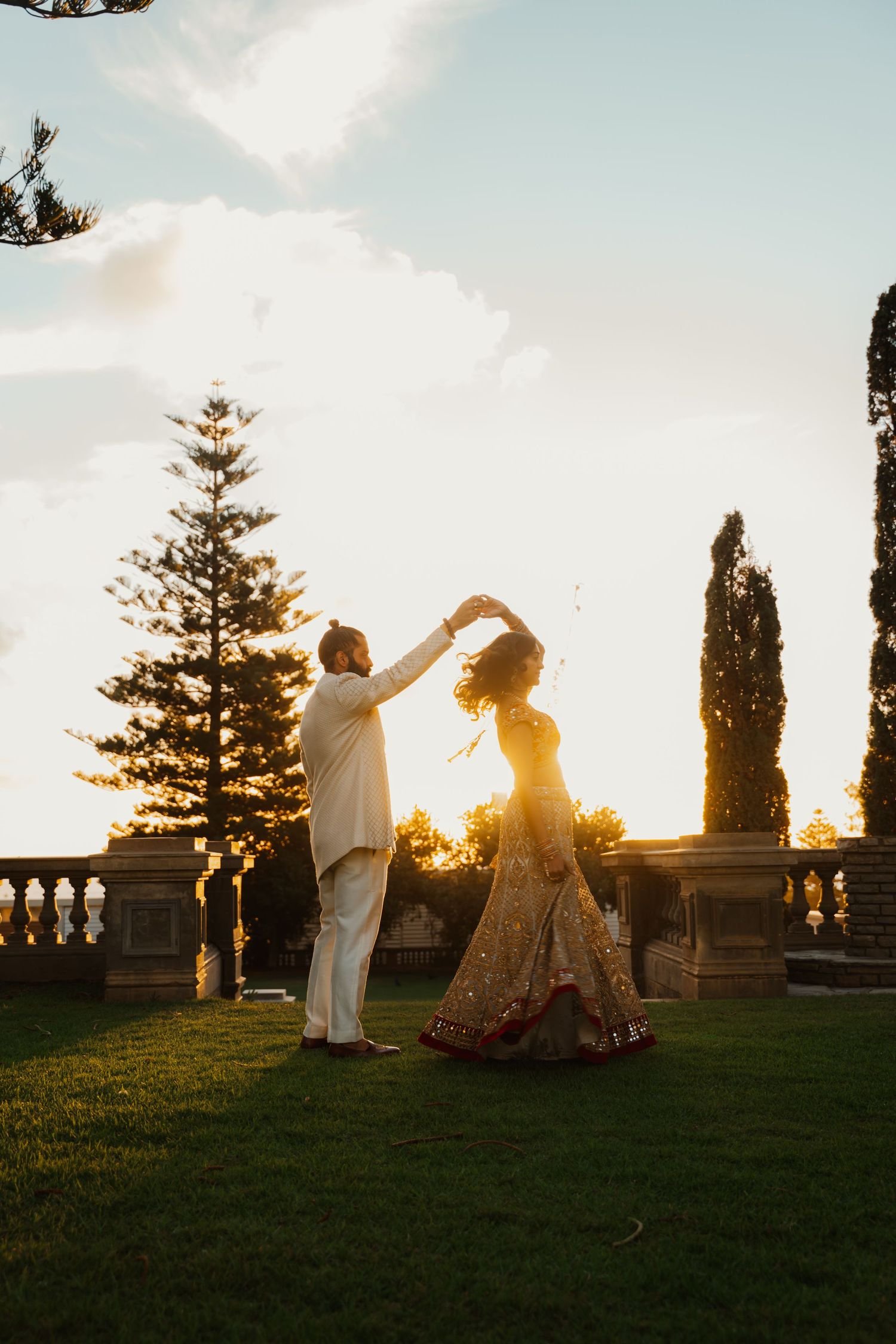 Indian groom twirls bride in gold lehenga on garden lawn during golden hour wedding portrait.