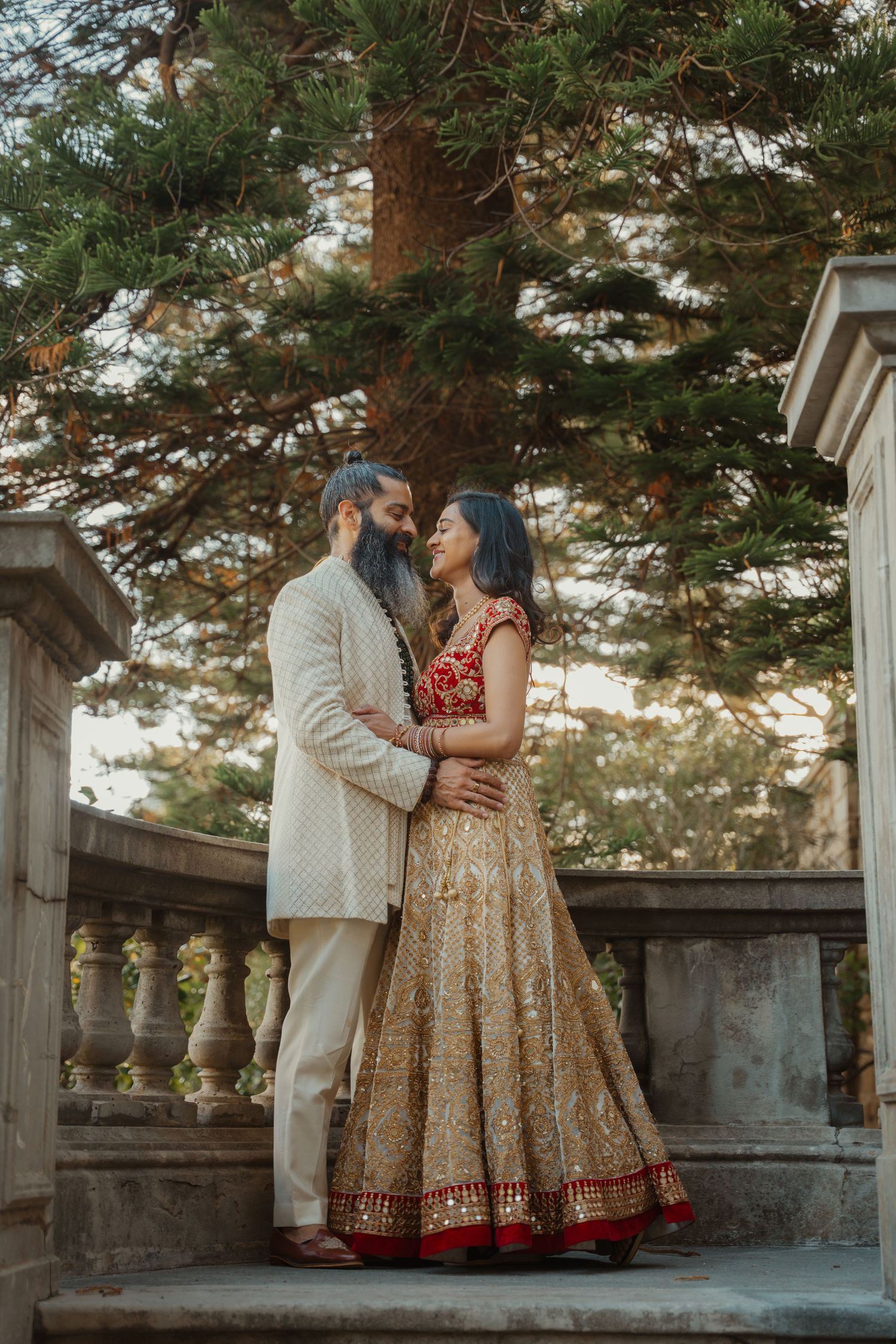 Indian couple in traditional wedding attire embrace on stone balcony surrounded by lush green trees.