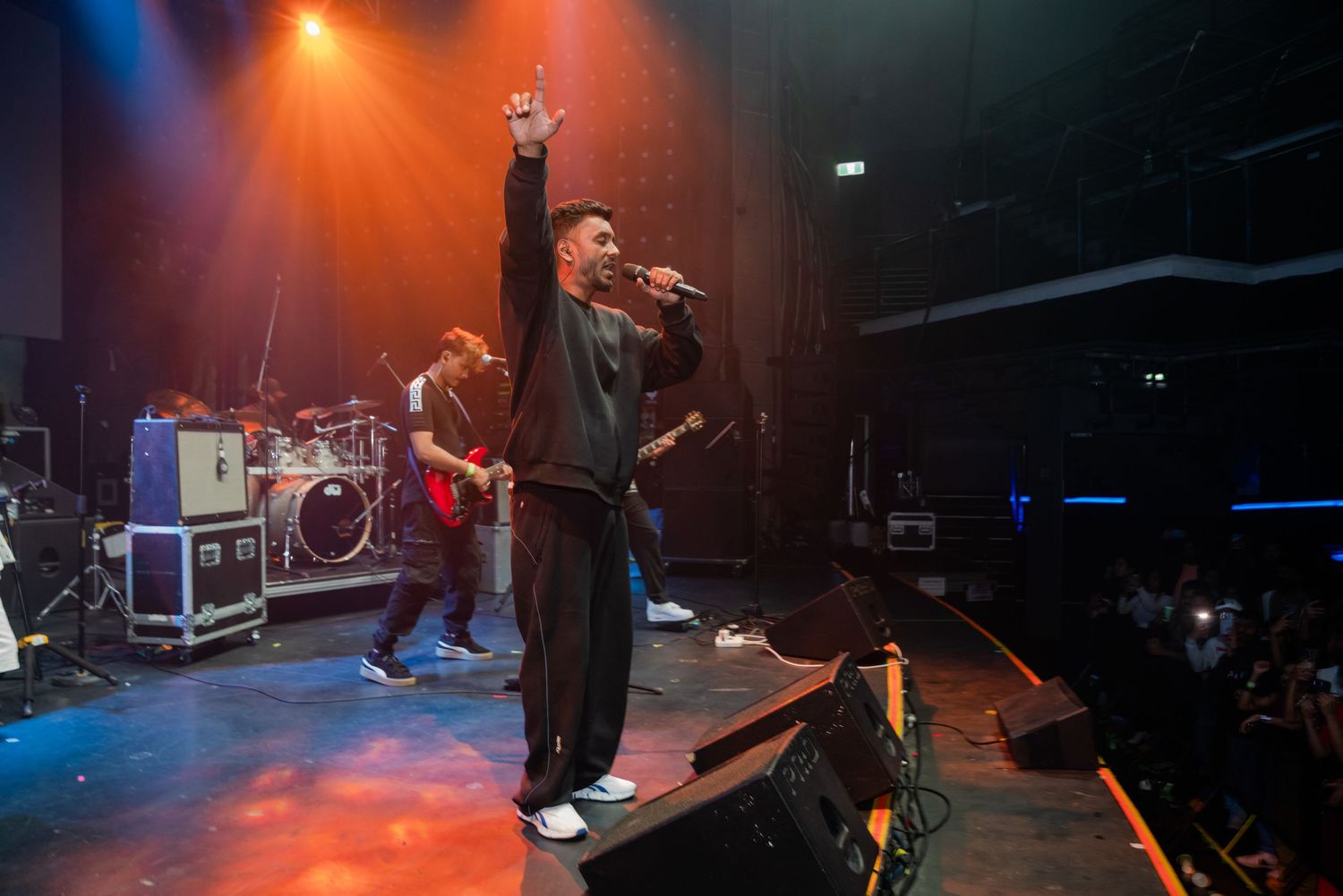 Singer raising hand on stage with guitarist and drummer performing behind him.