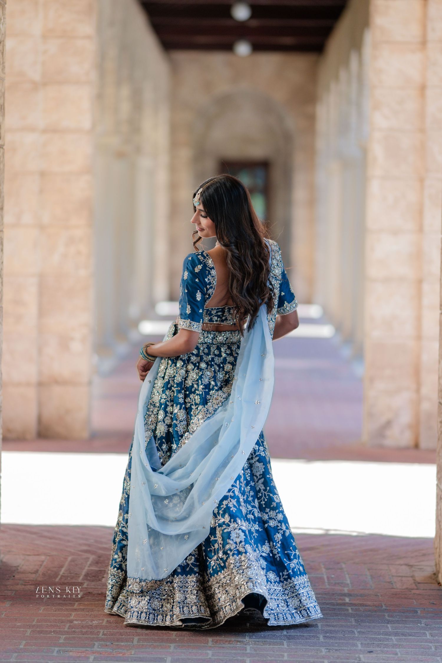 A woman in a blue floral lehenga with light blue dupatta poses in an elegant archway corridor.