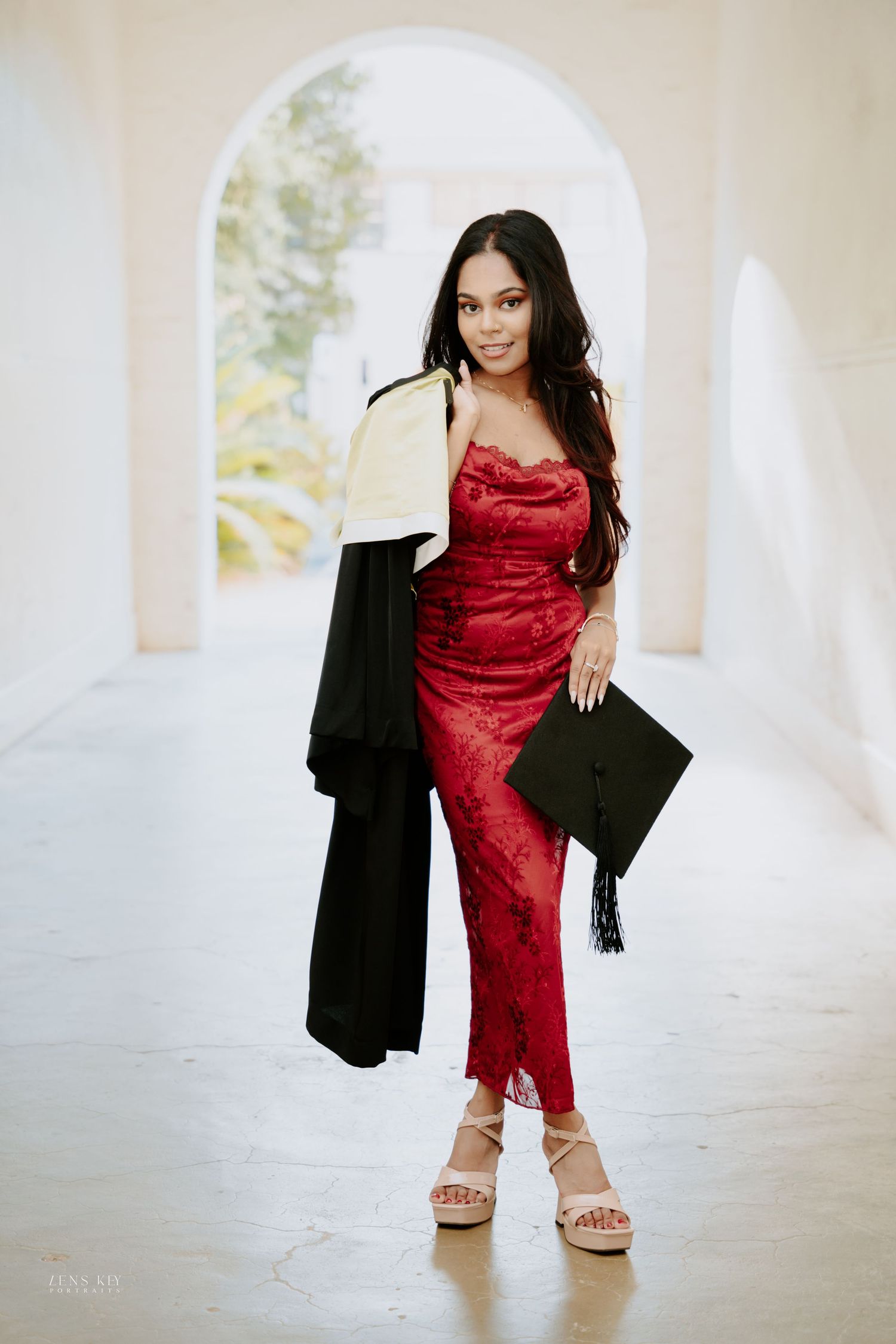 Young woman in red lace dress holding graduation cap and gown in elegant archway corridor.