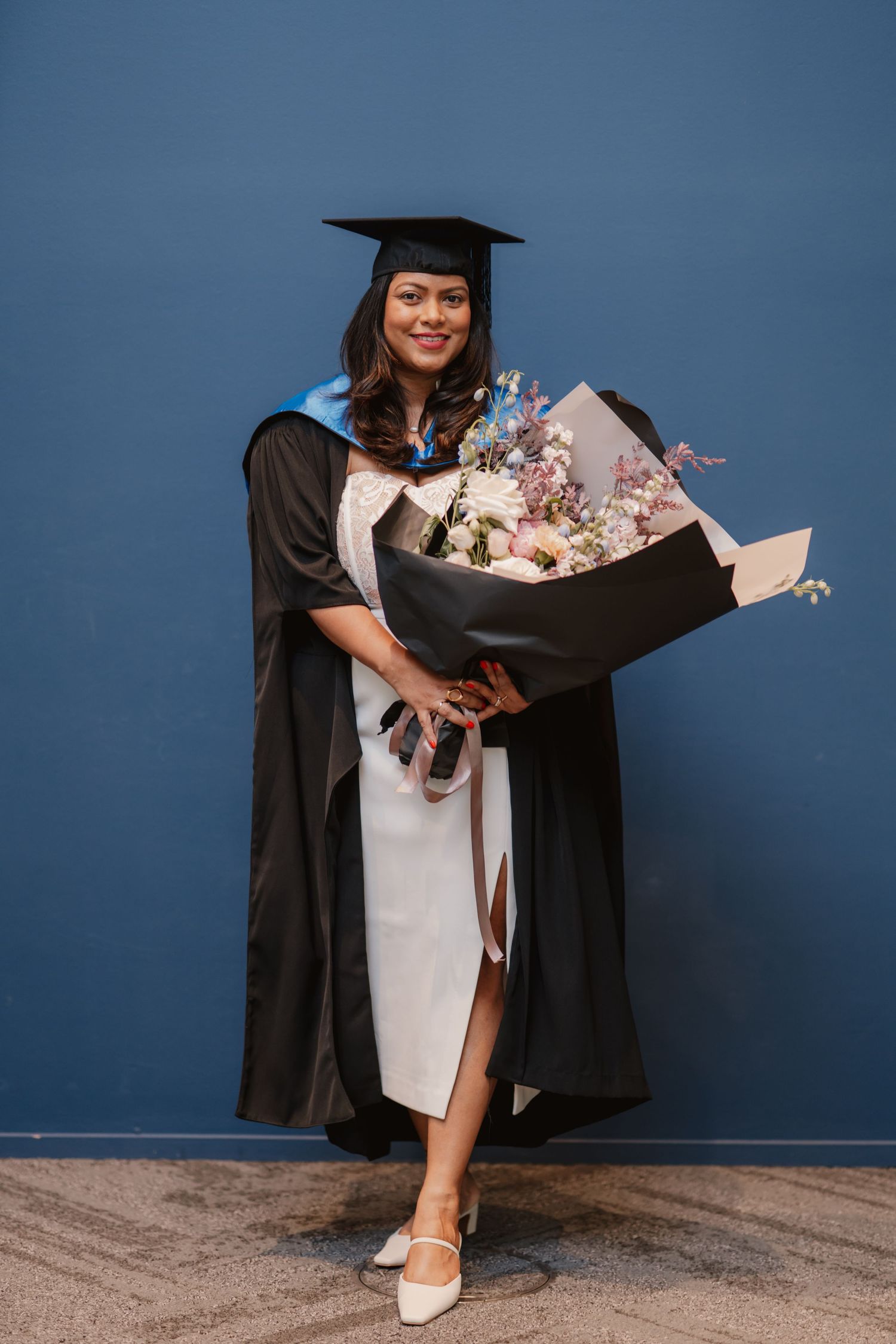 Joyful graduate in black cap and gown holds large floral bouquet against blue wall.